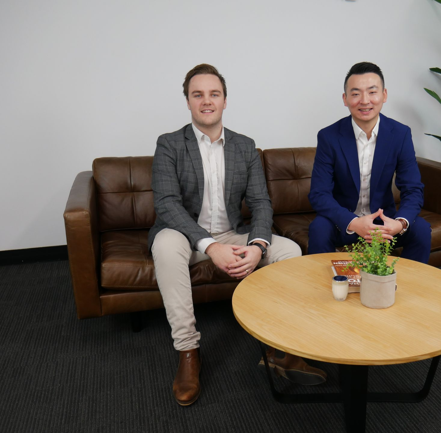 Two men in business attire sit on a leather couch, facing forward. A table with a plant sits in front of them.