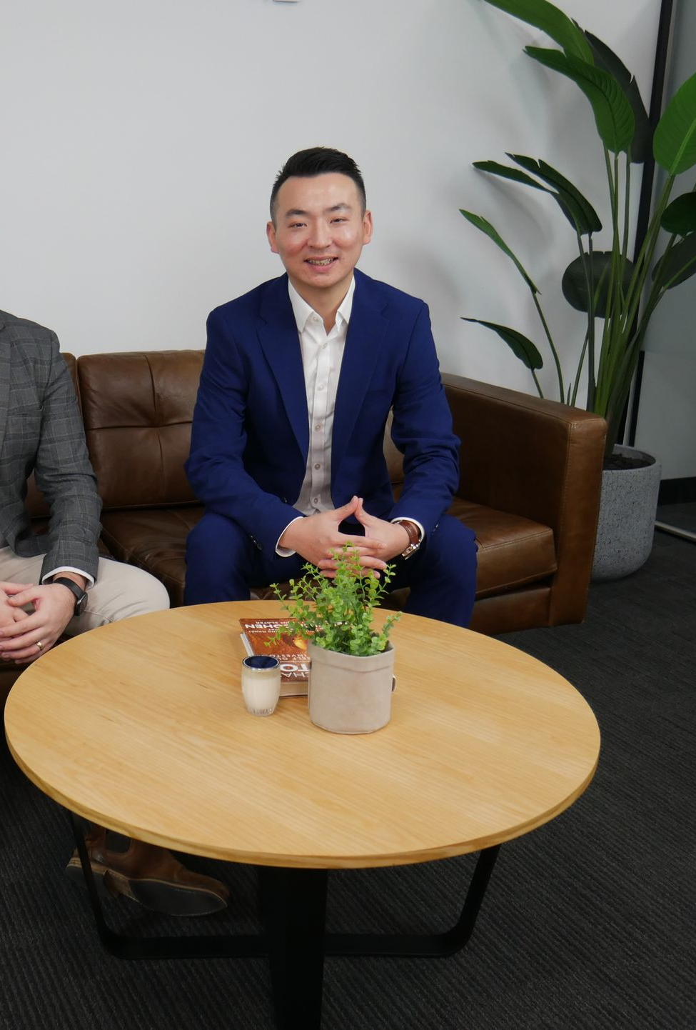 Man in blue suit sits on couch, smiling at the camera. A round table and plants are in front.