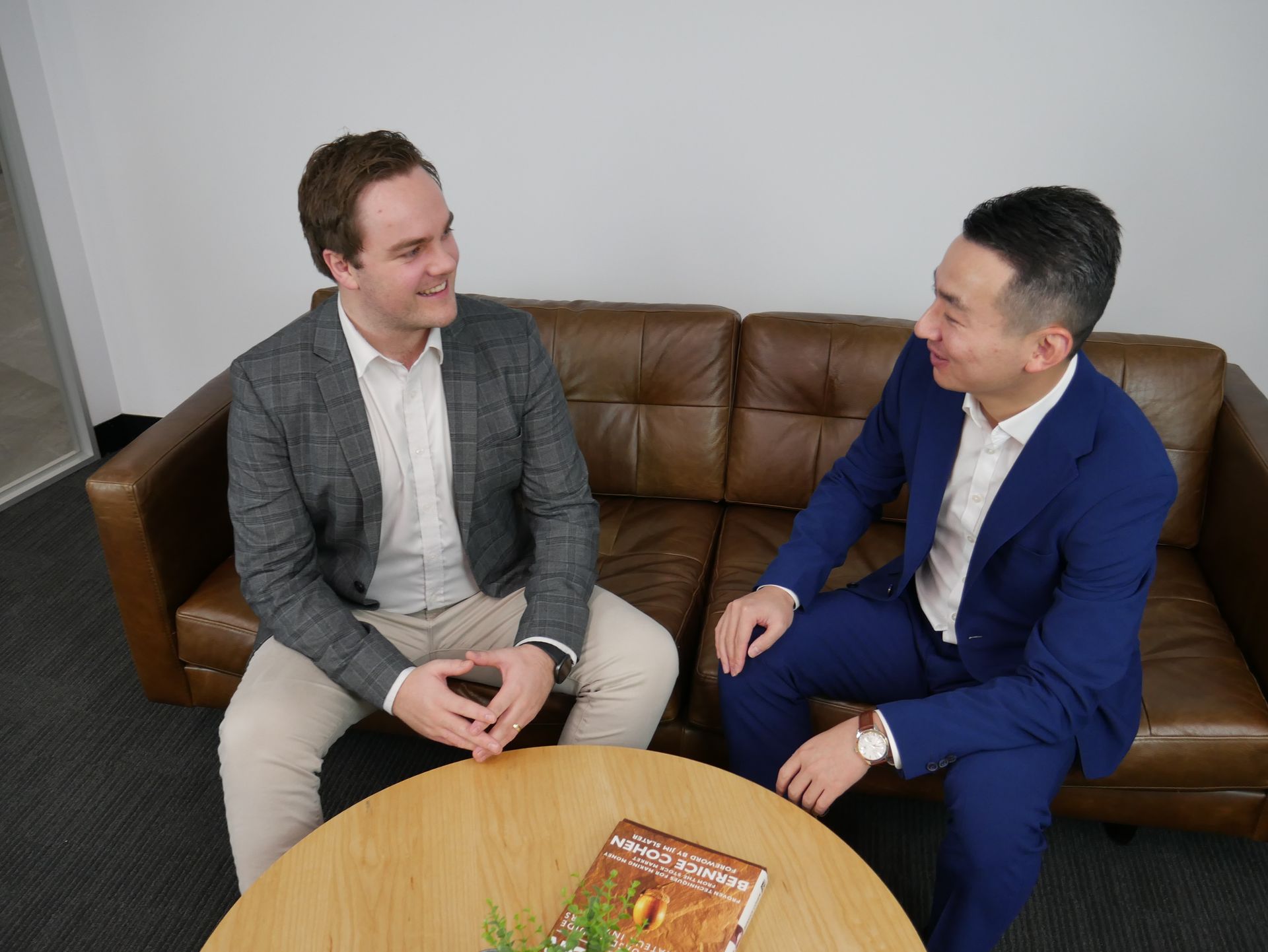 Two men in suits sit on a brown couch, engaged in a conversation at a table in a modern office setting.