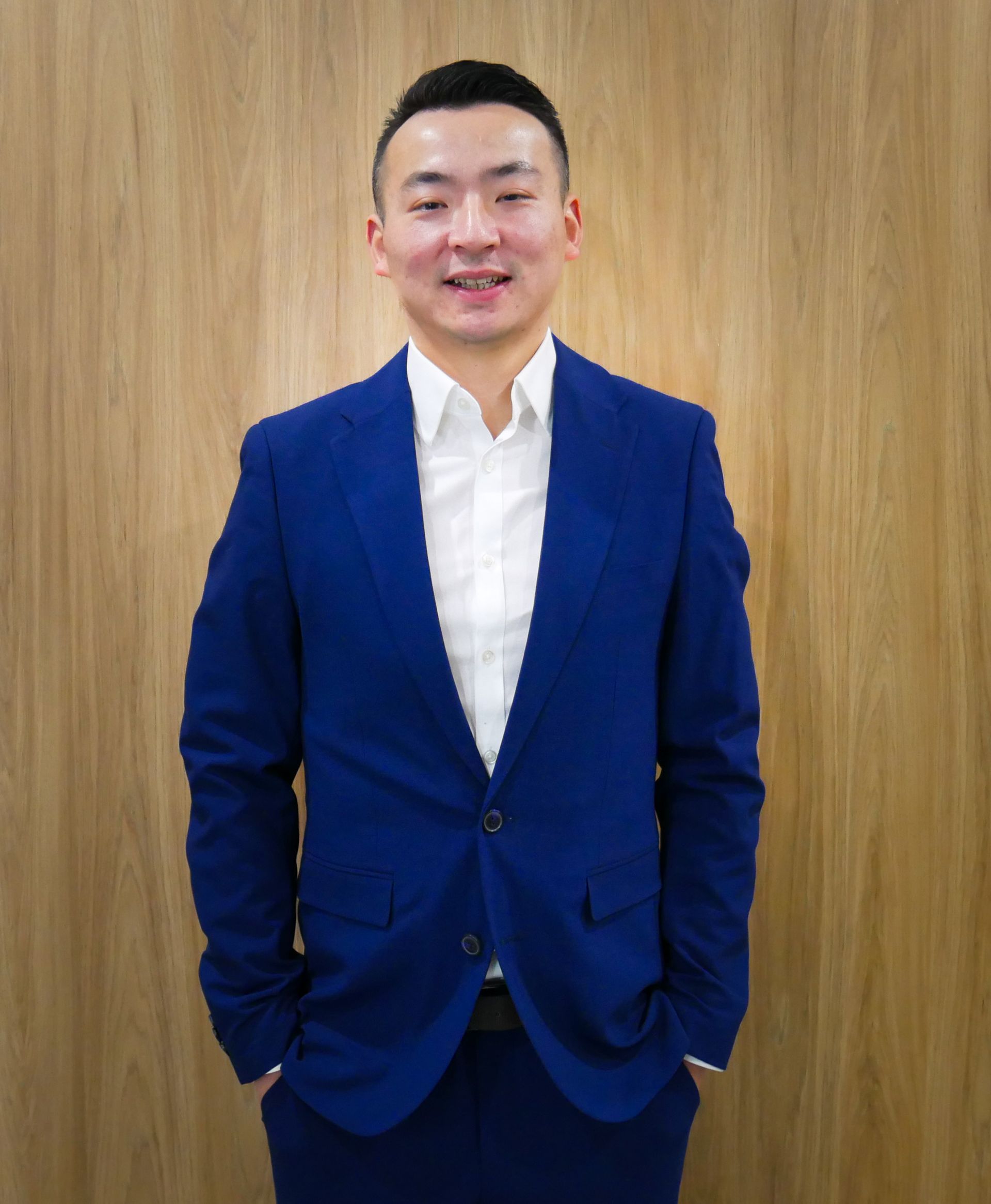Man in blue suit, hands in pockets, smiling. Standing in front of a wood-paneled wall.