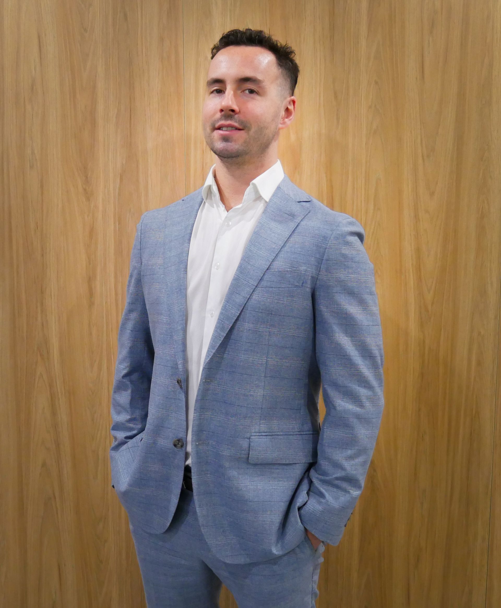 Man in blue suit and white shirt poses with hands in pockets, against a wood panel backdrop.