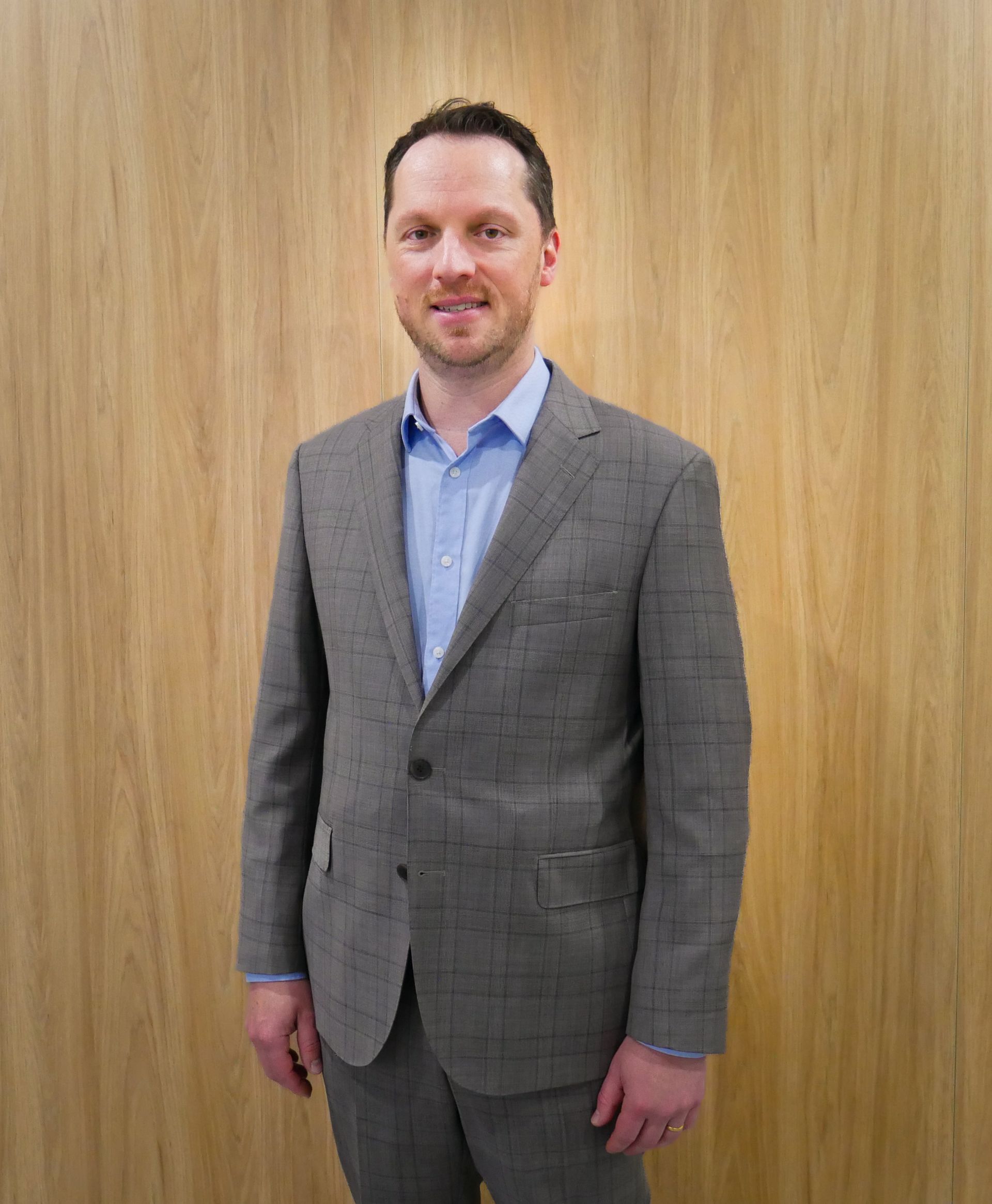 Man in a gray suit and blue shirt smiles, standing against a wood-paneled wall.