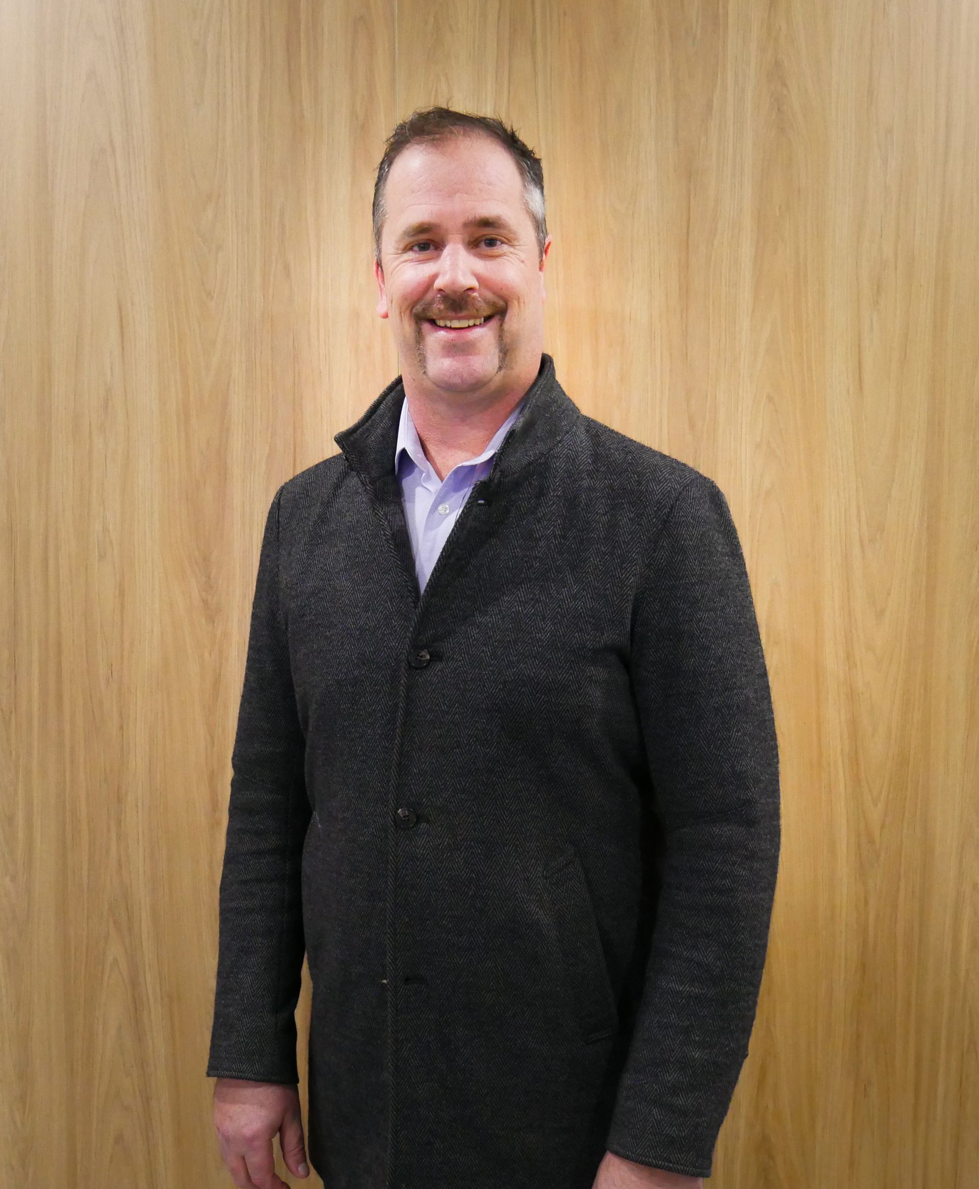 Man smiling in a dark gray sweater and blue shirt, standing in front of wooden panels.