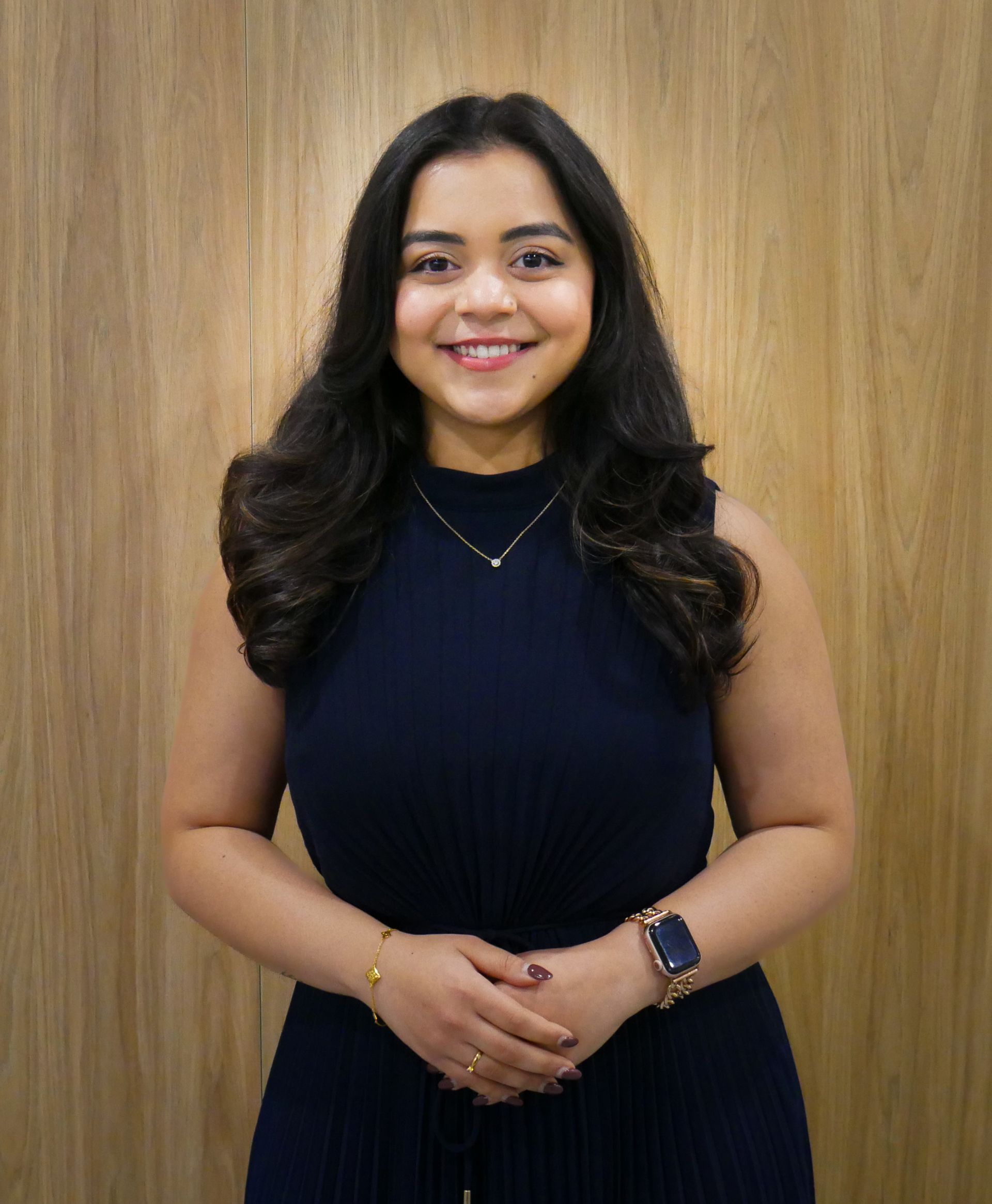 Woman smiling, arms crossed, in a navy blue sleeveless dress, gold jewelry, against a wood-paneled wall.