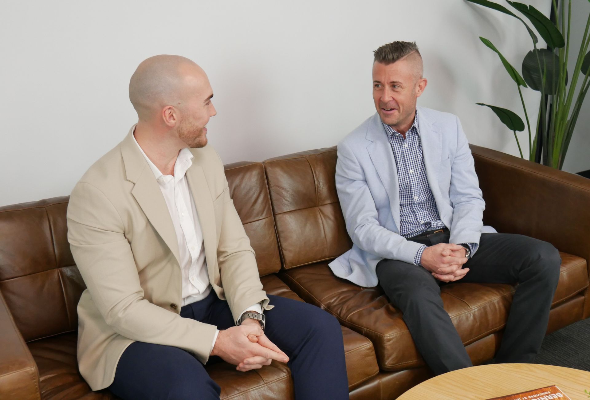 Two men in blazers seated on a brown leather couch, conversing in an office setting.