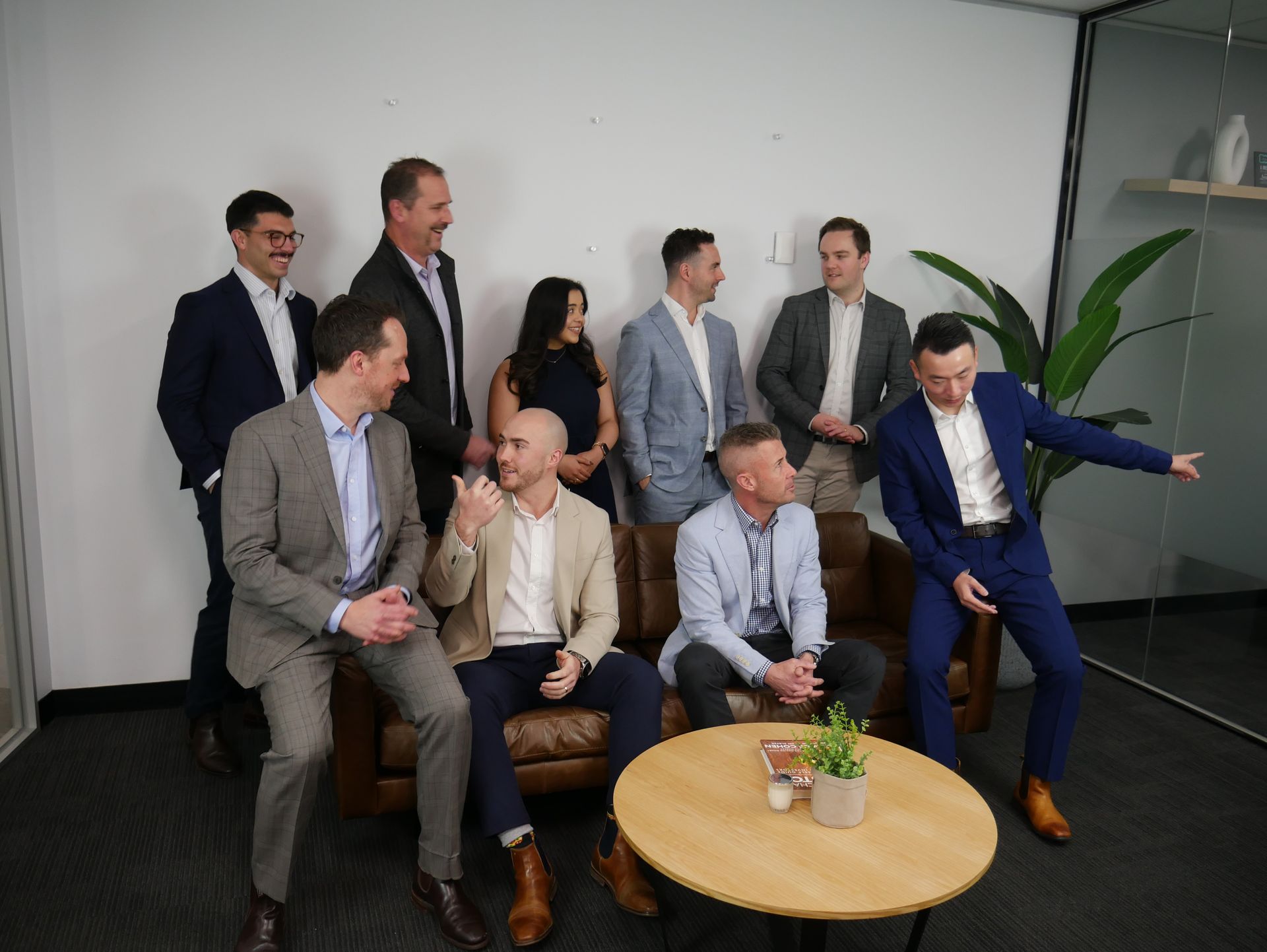 Group of suited people in an office, some standing, some seated on a couch, one pointing.