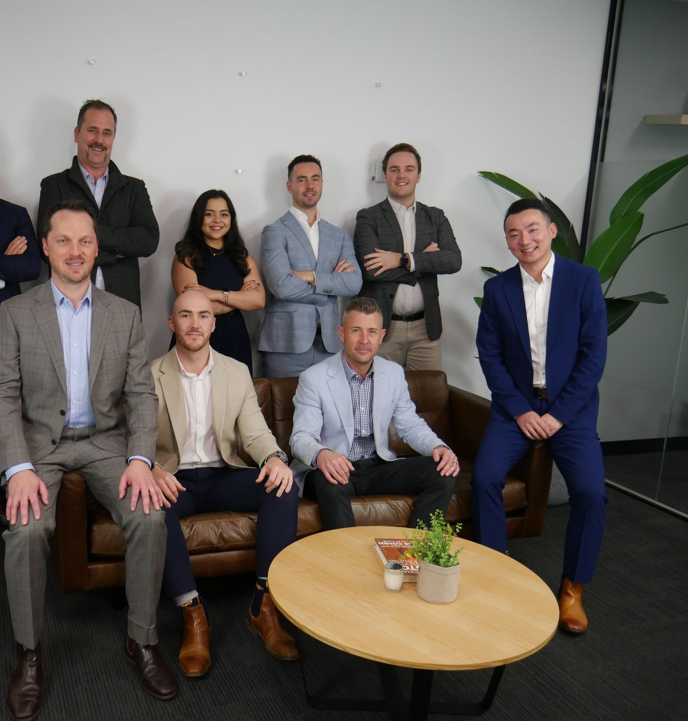 Group of business professionals in suits posing in an office. Some seated, others standing, smiling at camera.