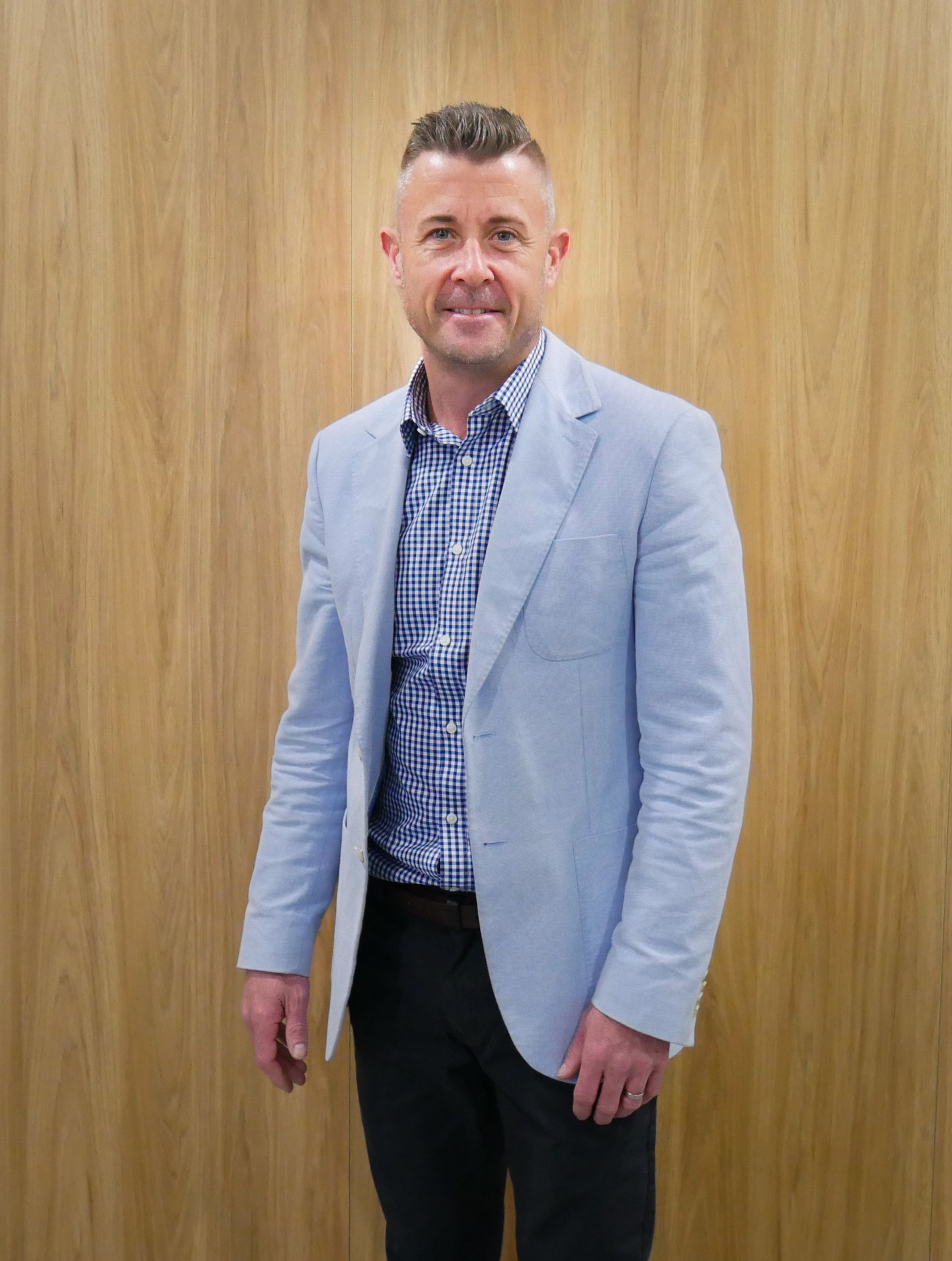 Man in light blue blazer and patterned shirt stands in front of a wood-paneled wall.