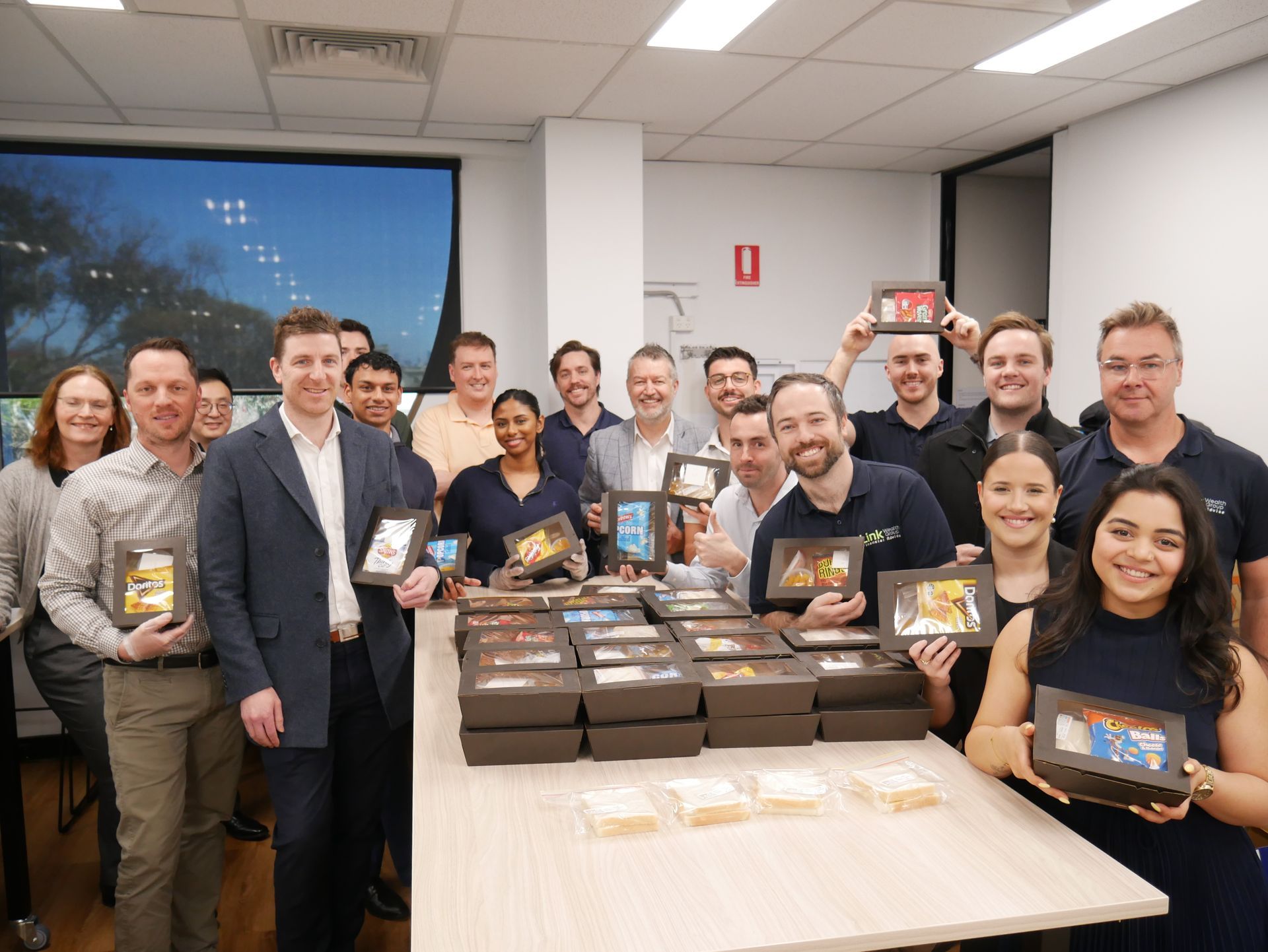 Group of people in an office holding gift boxes, smiling, posing near a table of treats.
