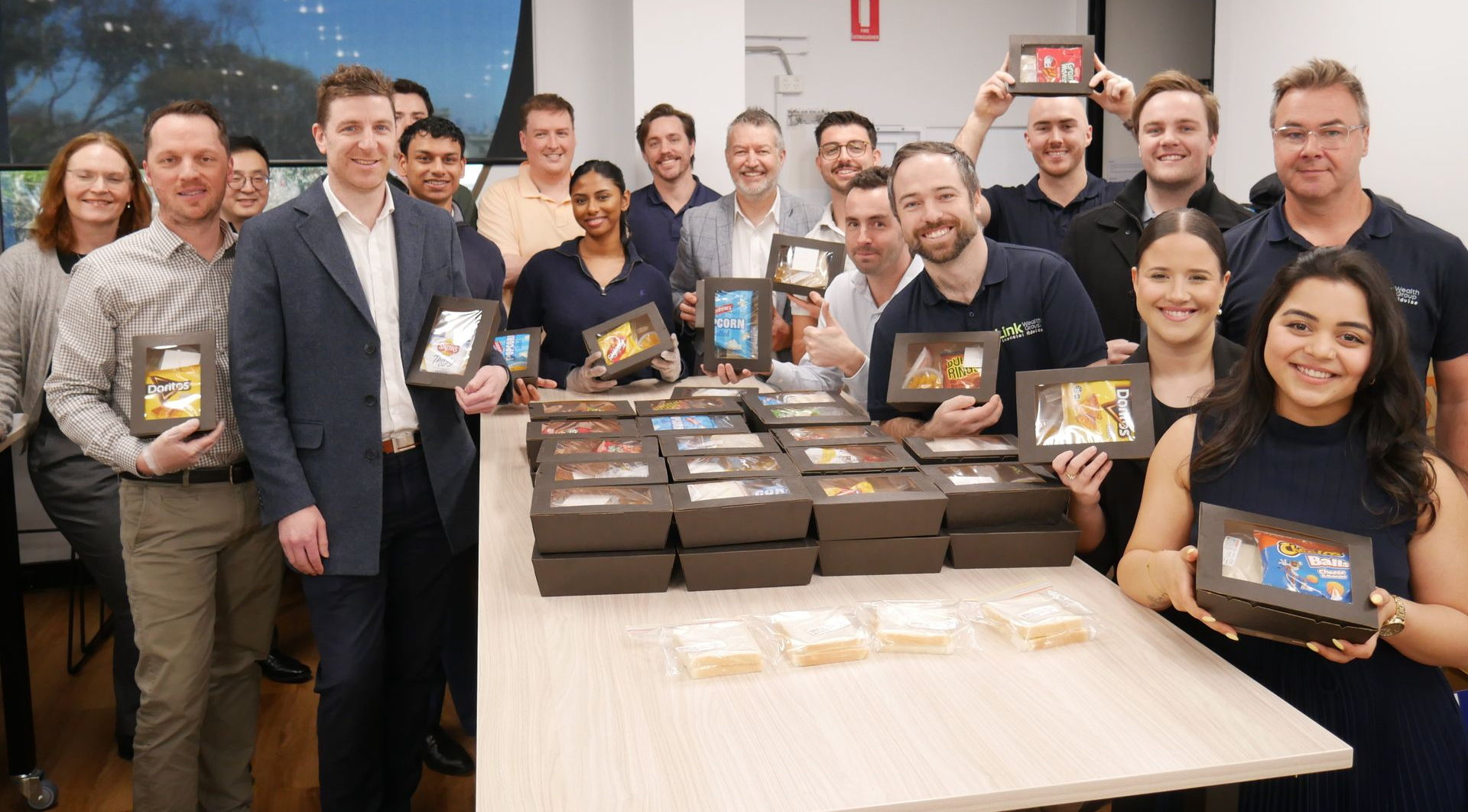 Group of people, mostly in business attire, hold up gift boxes, smiling in an office setting.