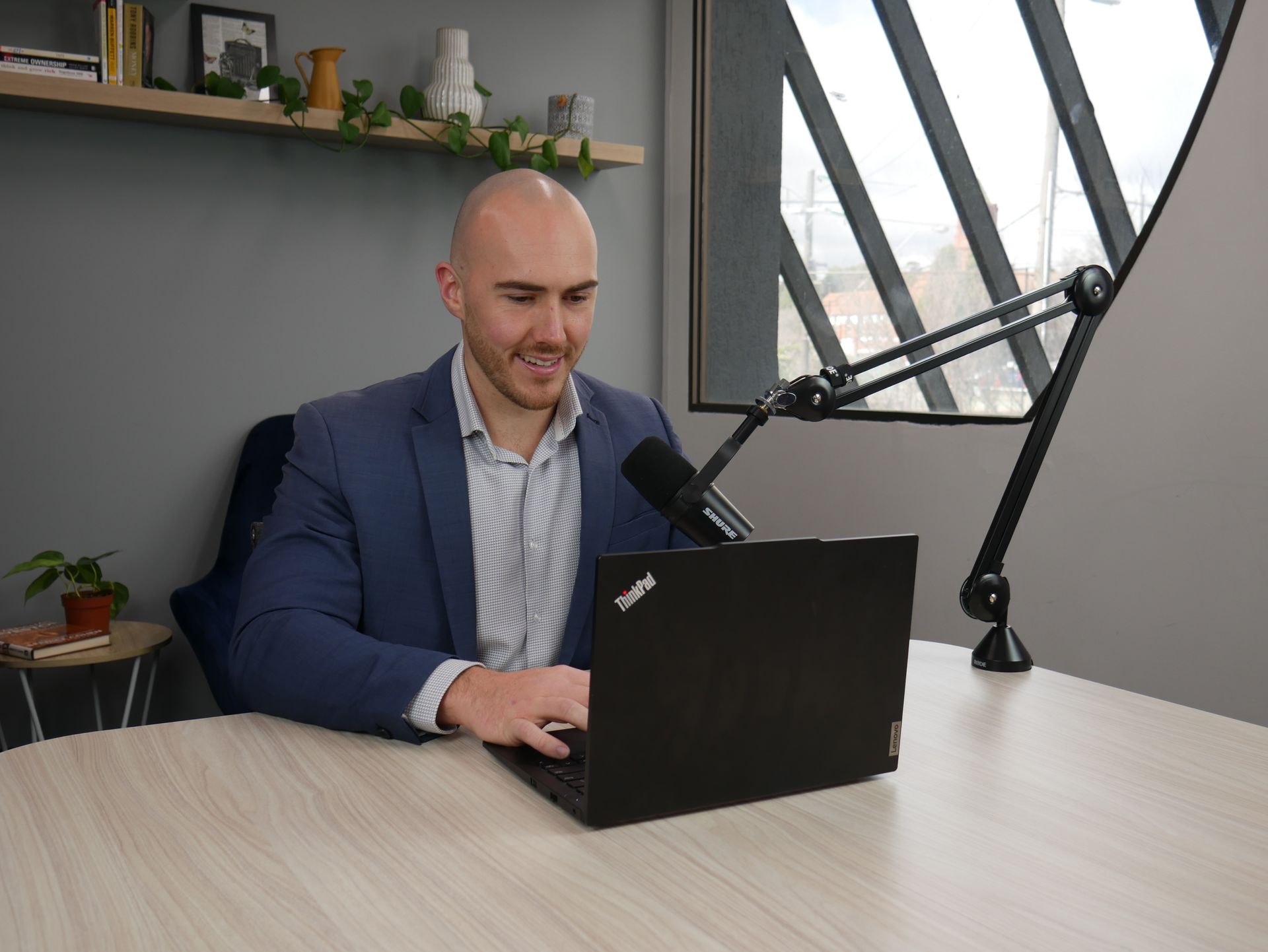 Man in a blue suit, typing on a laptop at a desk with a microphone, grey wall background.