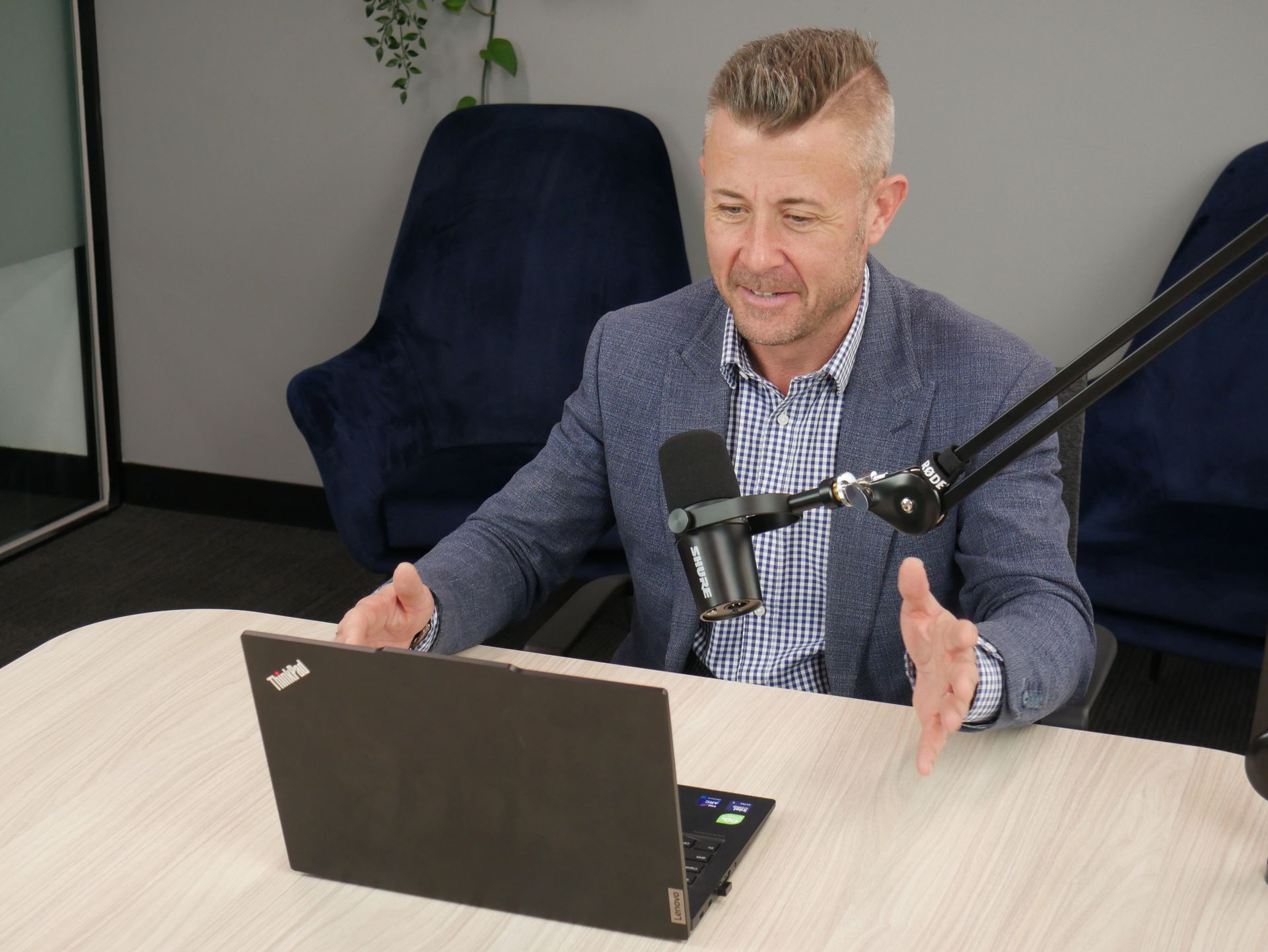 Man in blue blazer speaks at desk, gesturing. Laptop and microphone are present.