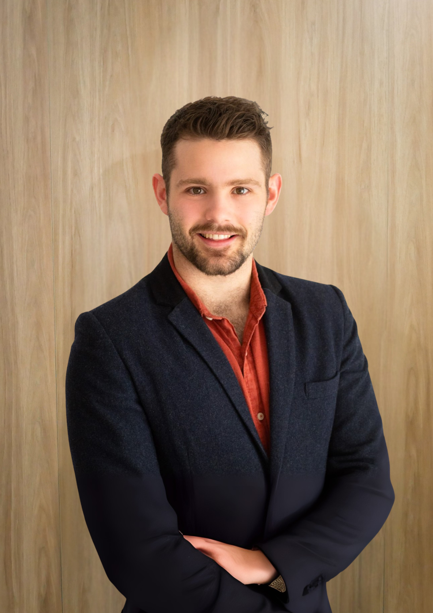 Man with a beard and a smile, wearing a navy blazer and red shirt, arms crossed, standing in front of a wood-paneled wall.