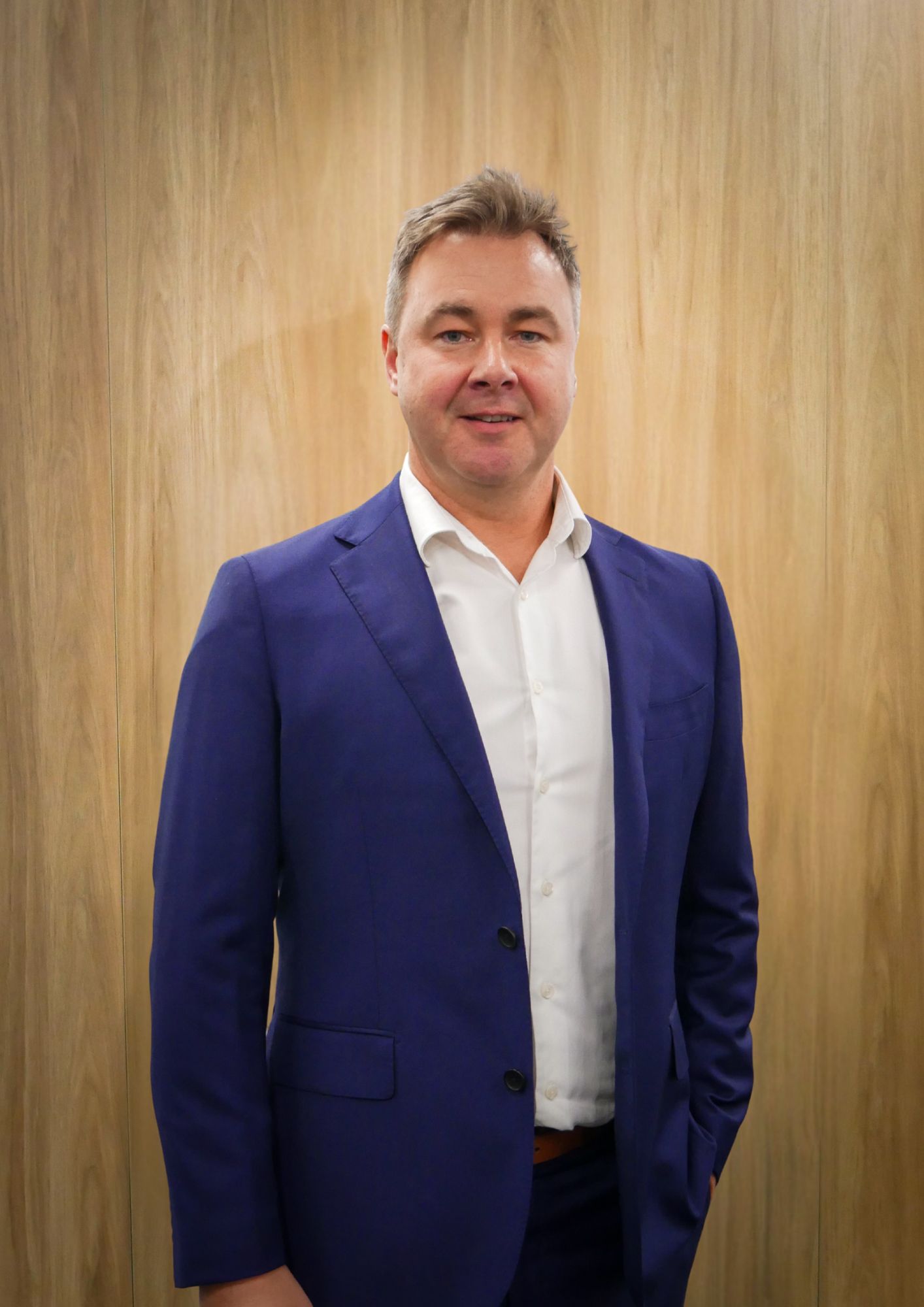 Man in a blue suit and white shirt smiles, standing against a wood-paneled wall.