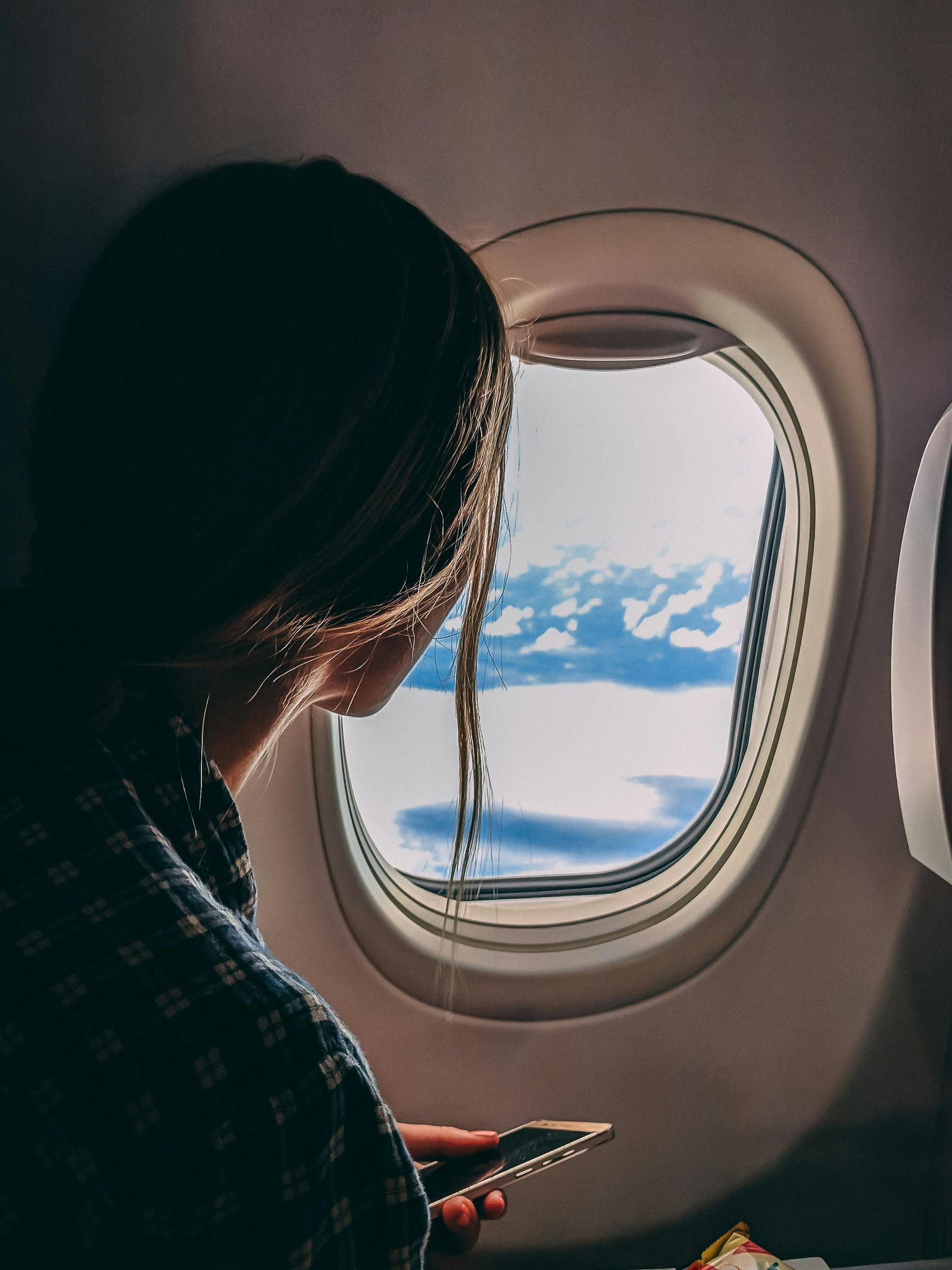 Woman in plaid shirt looking out airplane window at clouds, holding a phone.