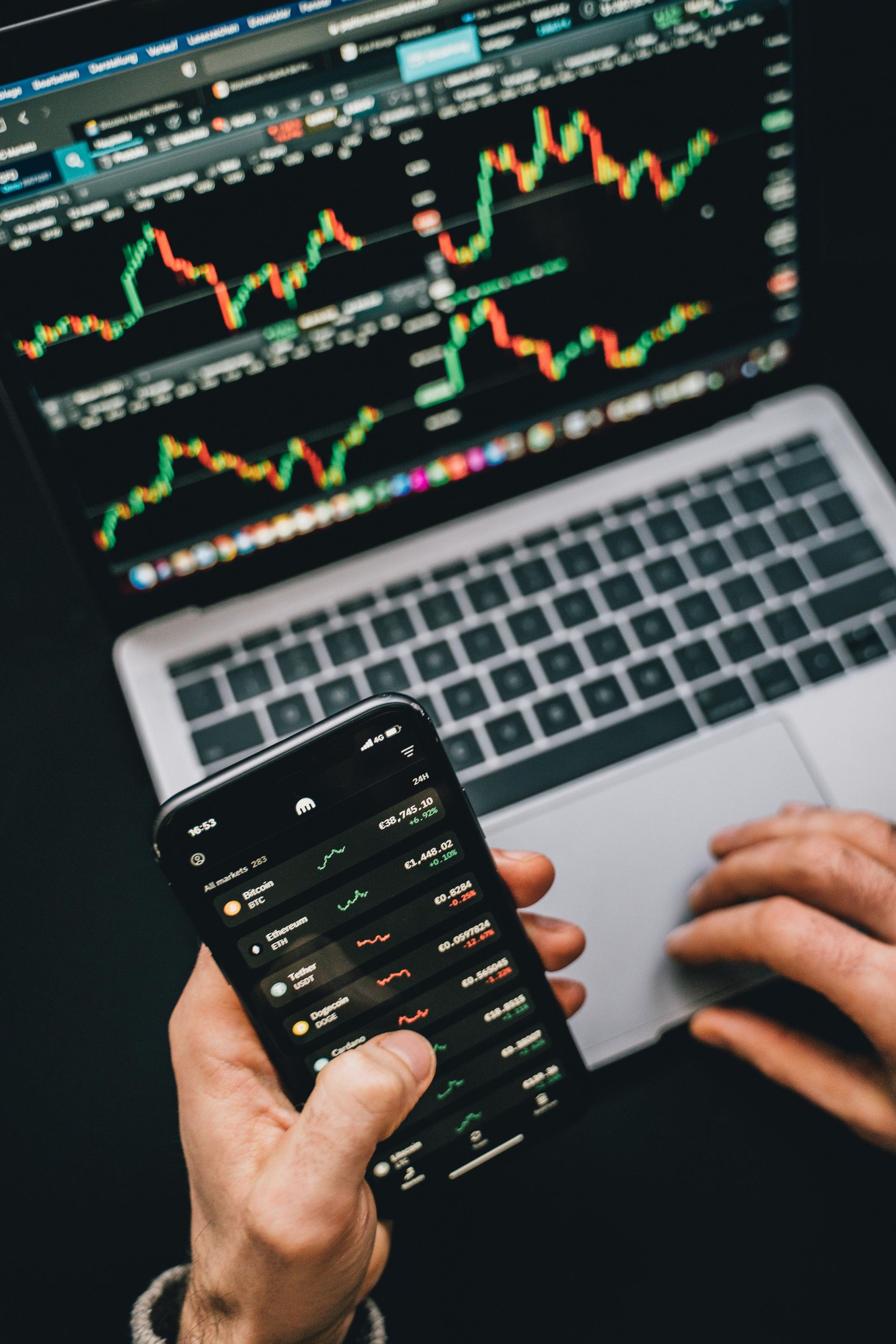 Person holding a smartphone and using a laptop to monitor stock market charts on a dark surface.