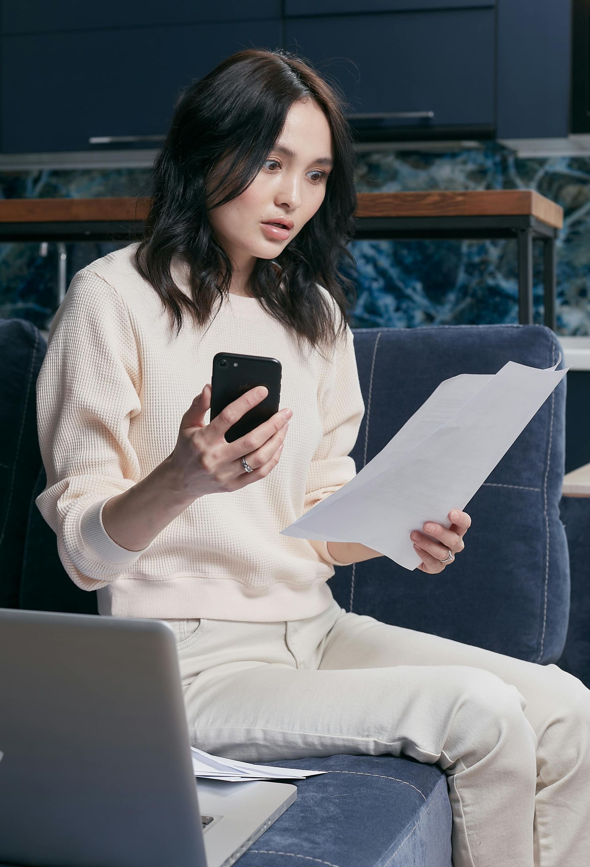 Woman looking at phone and paperwork, seated on a couch. Laptop nearby. Neutral clothing, concerned expression.