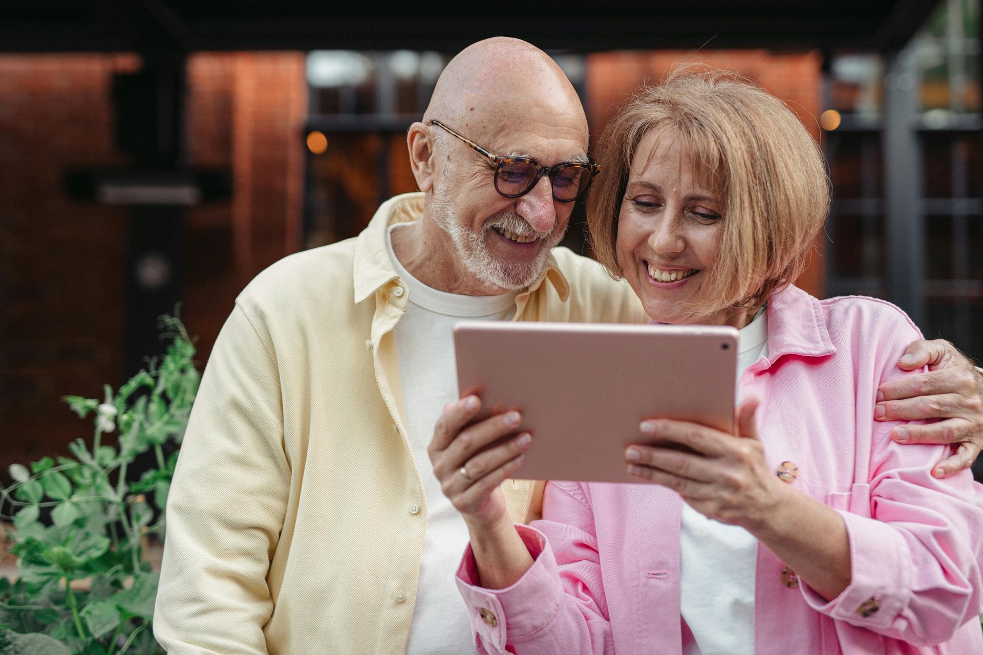 Smiling elderly couple looking at a tablet; man has arm around woman outside a brick building.