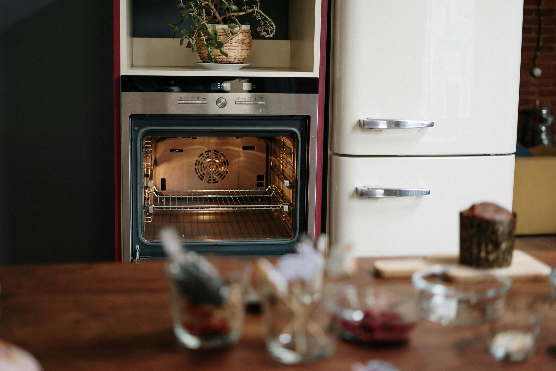 Oven with open door, plant above, and retro refrigerator. Table has glasses and a muffin.