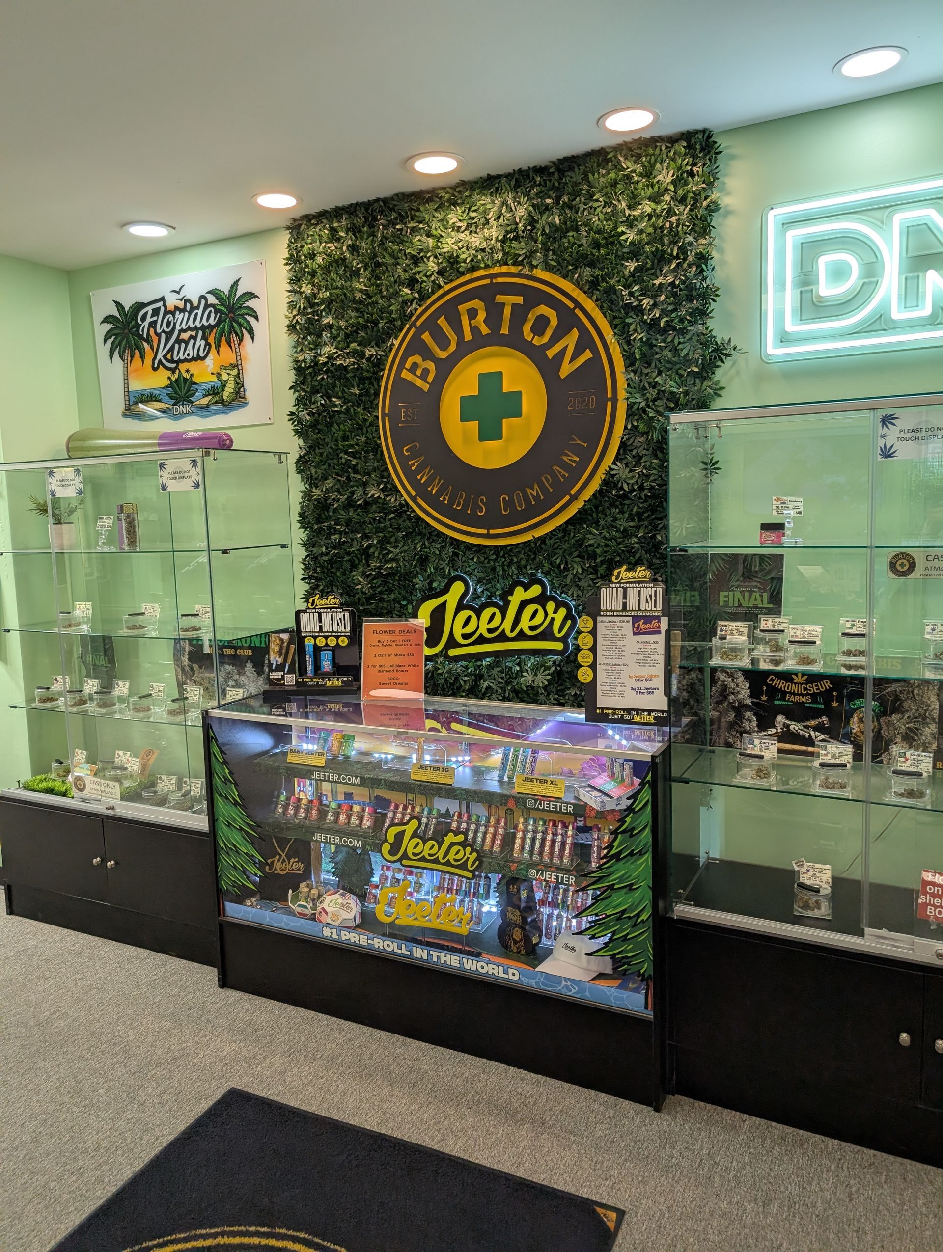 Interior of a cannabis dispensary. Counter with products, glass display cases, Burton logo, and neon sign.