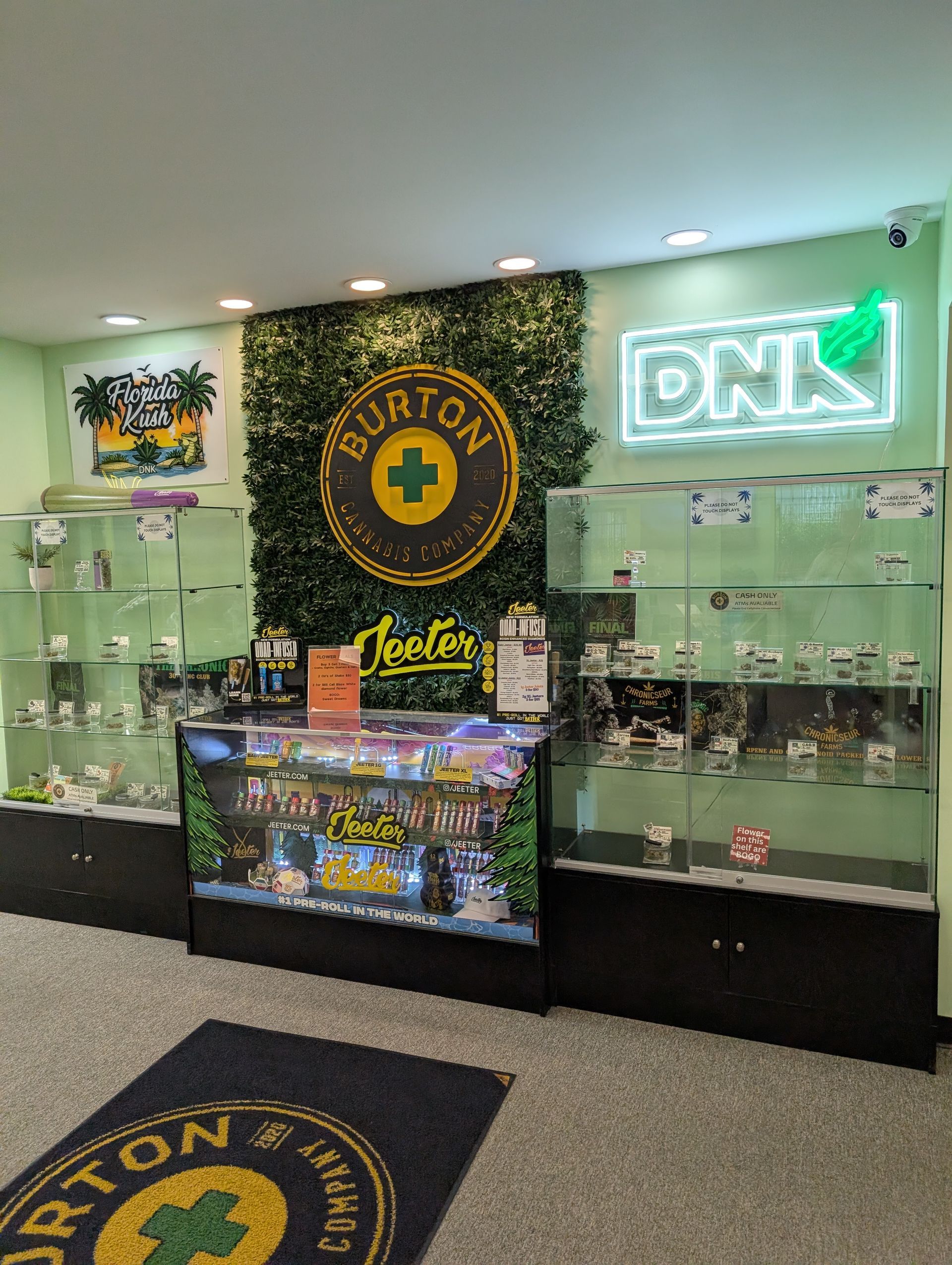 Interior of a dispensary with display cases, neon sign, and Burton logo on a green wall.