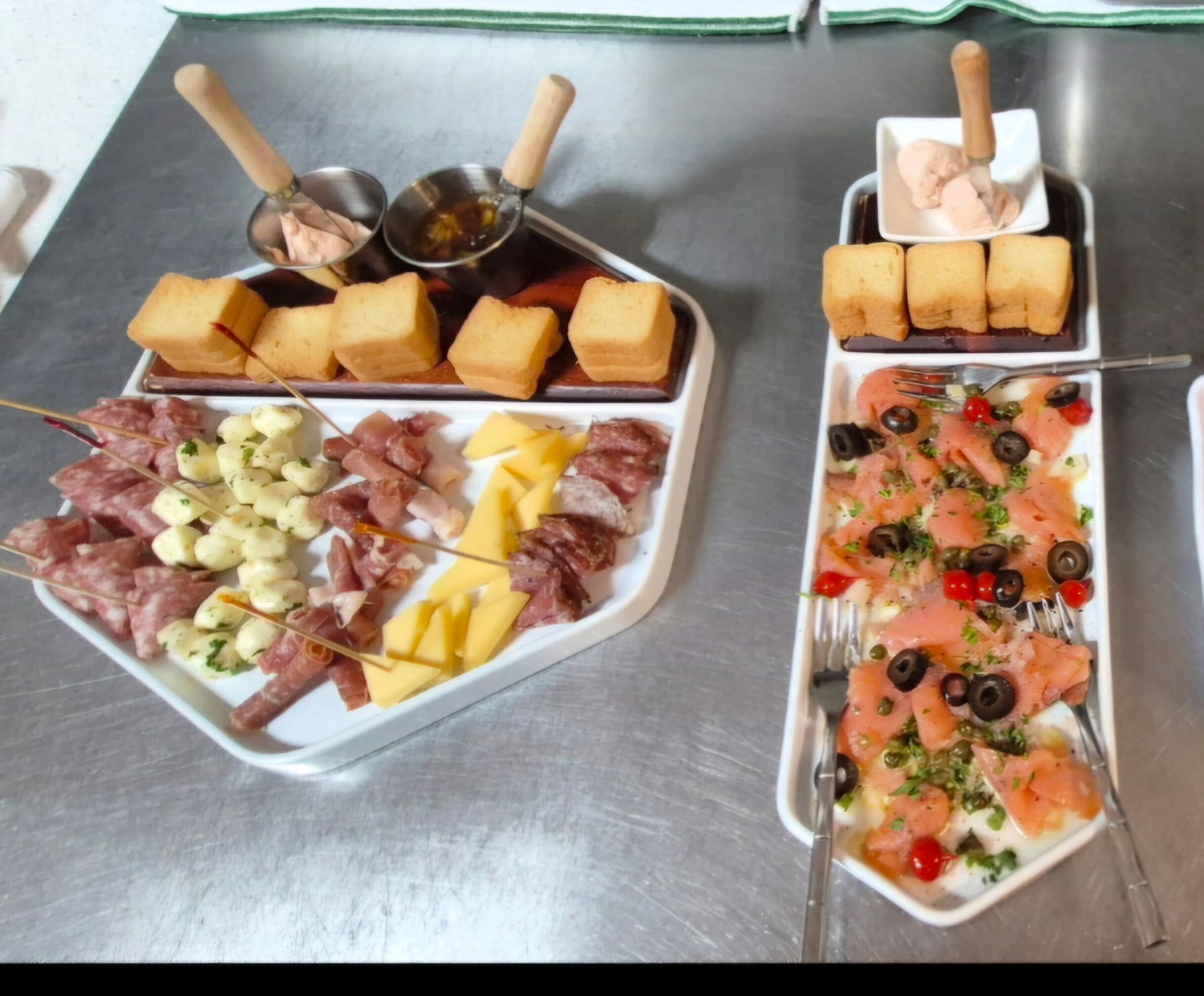 Two appetizer platters with meats, cheeses, salmon, and bread, on a metal surface.