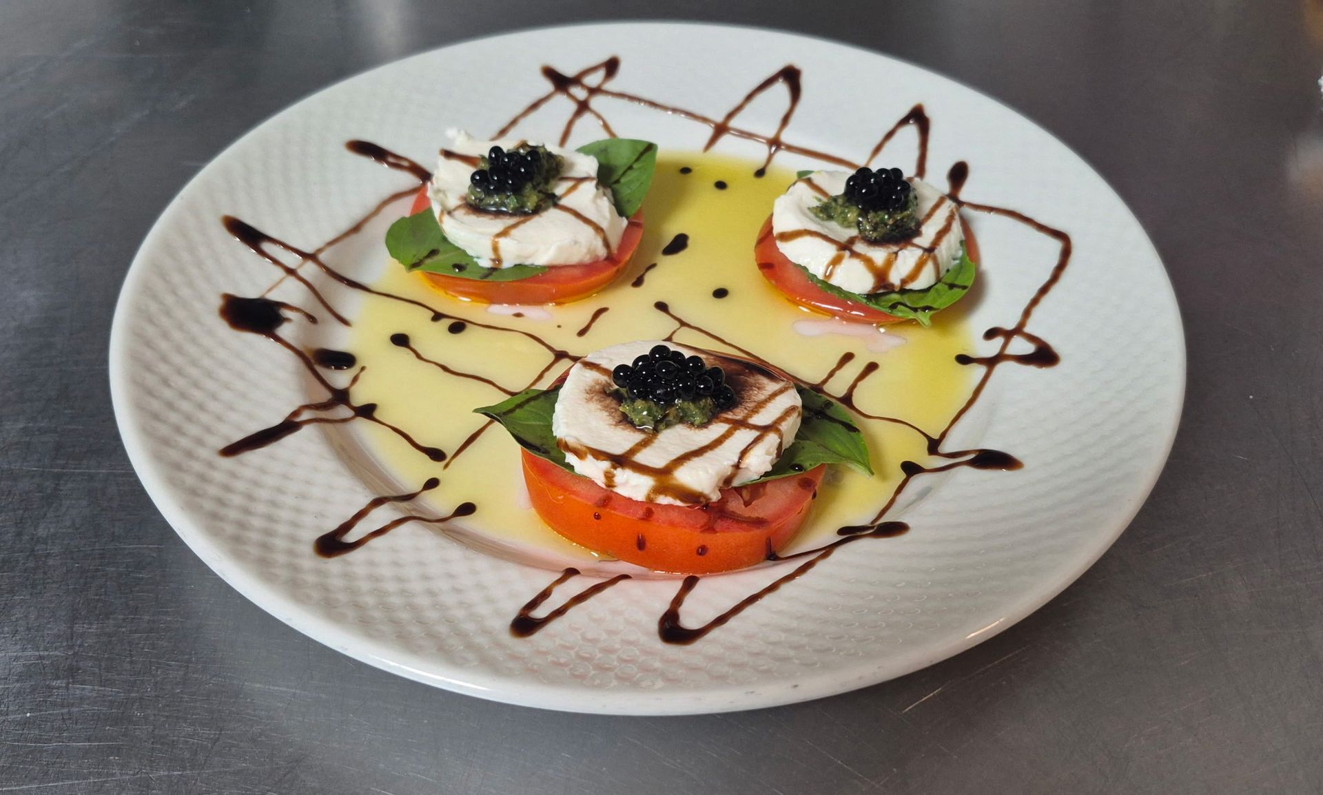 Caprese salad with tomato, mozzarella, basil, caviar, and balsamic glaze on a white plate.