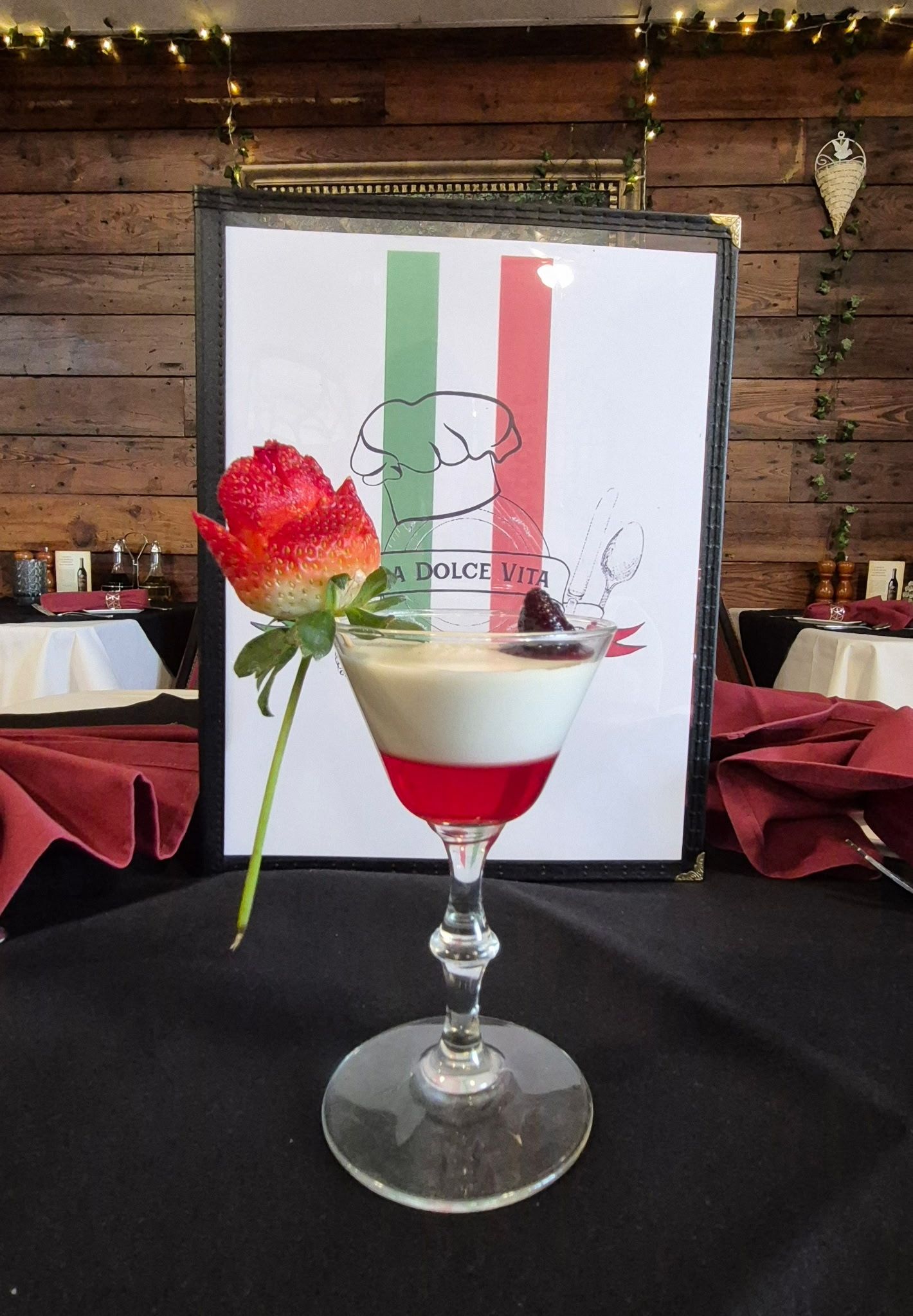 Dessert in a stemmed glass with red, white, and clear layers. A strawberry decorates the drink in front of Italian flag signage.
