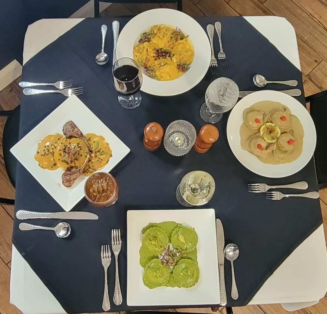 Table set for four with plates of pasta, sauces, silverware, glasses, and a bottle of wine on a blue tablecloth.