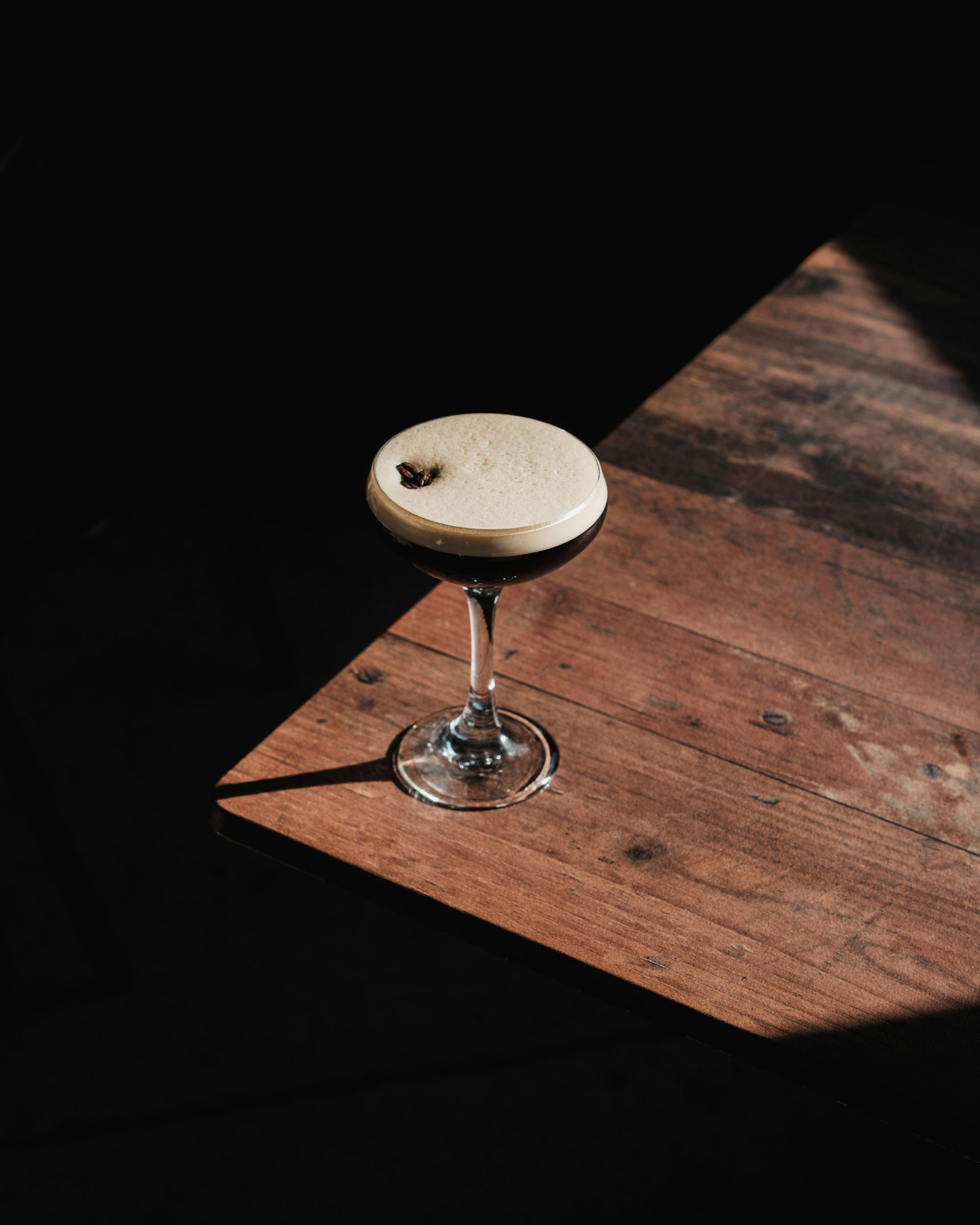 Espresso martini in a stemmed glass on a wooden surface, dark liquid topped with foamy cream, coffee bean garnish.