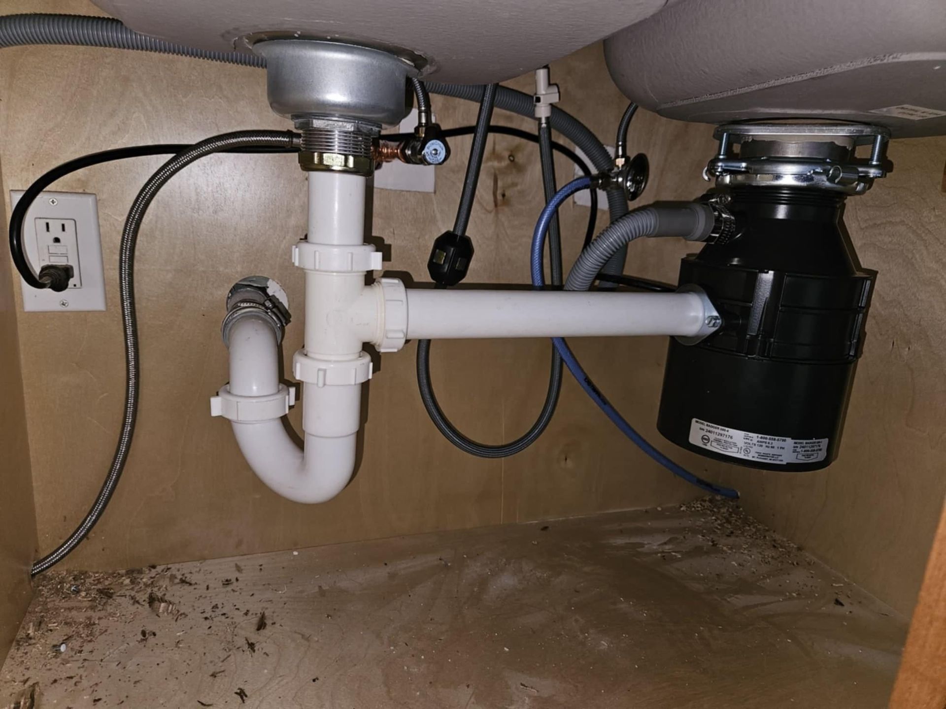 Under-sink plumbing with white pipes, a garbage disposal, and a sink drain. There's also an electrical outlet on the wall.