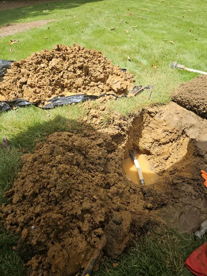 Hole dug in lawn reveals a broken pipe with muddy water, piles of brown dirt nearby.