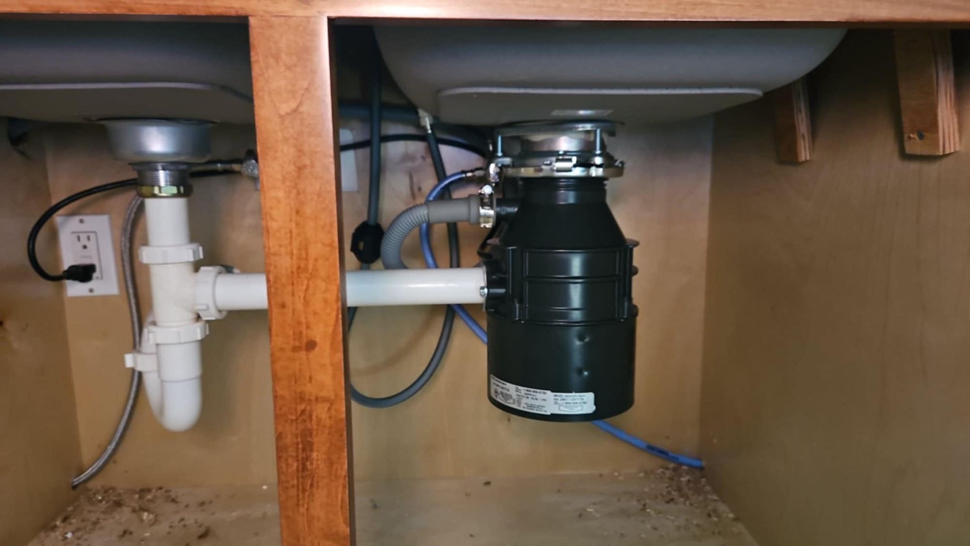 Under-sink plumbing with a garbage disposal; white pipes, black disposal, and electrical outlet inside a cabinet.