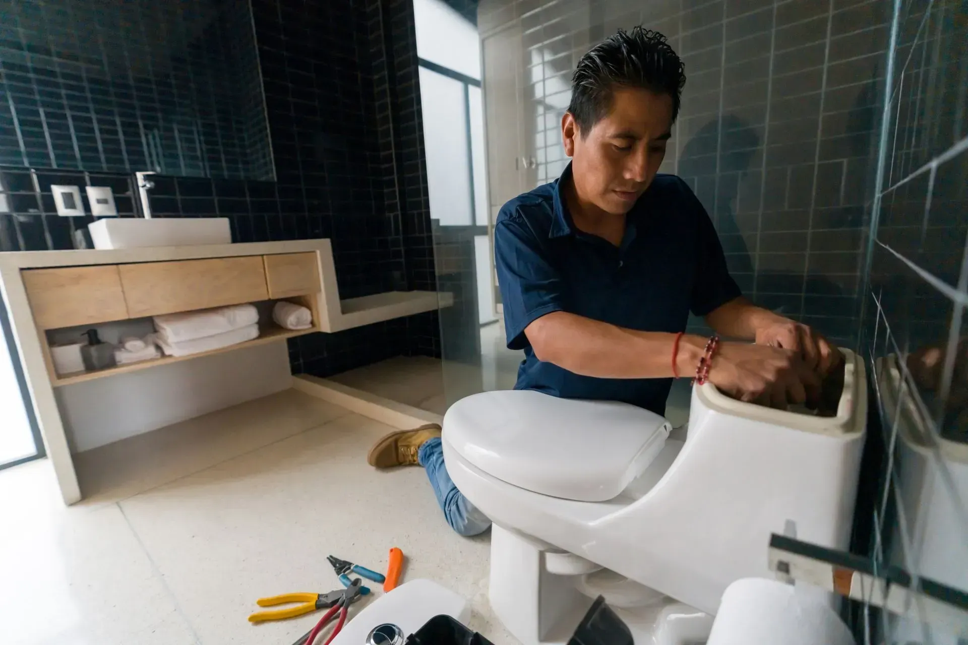Man in blue shirt repairing a white toilet in a modern bathroom. Tools scattered nearby.