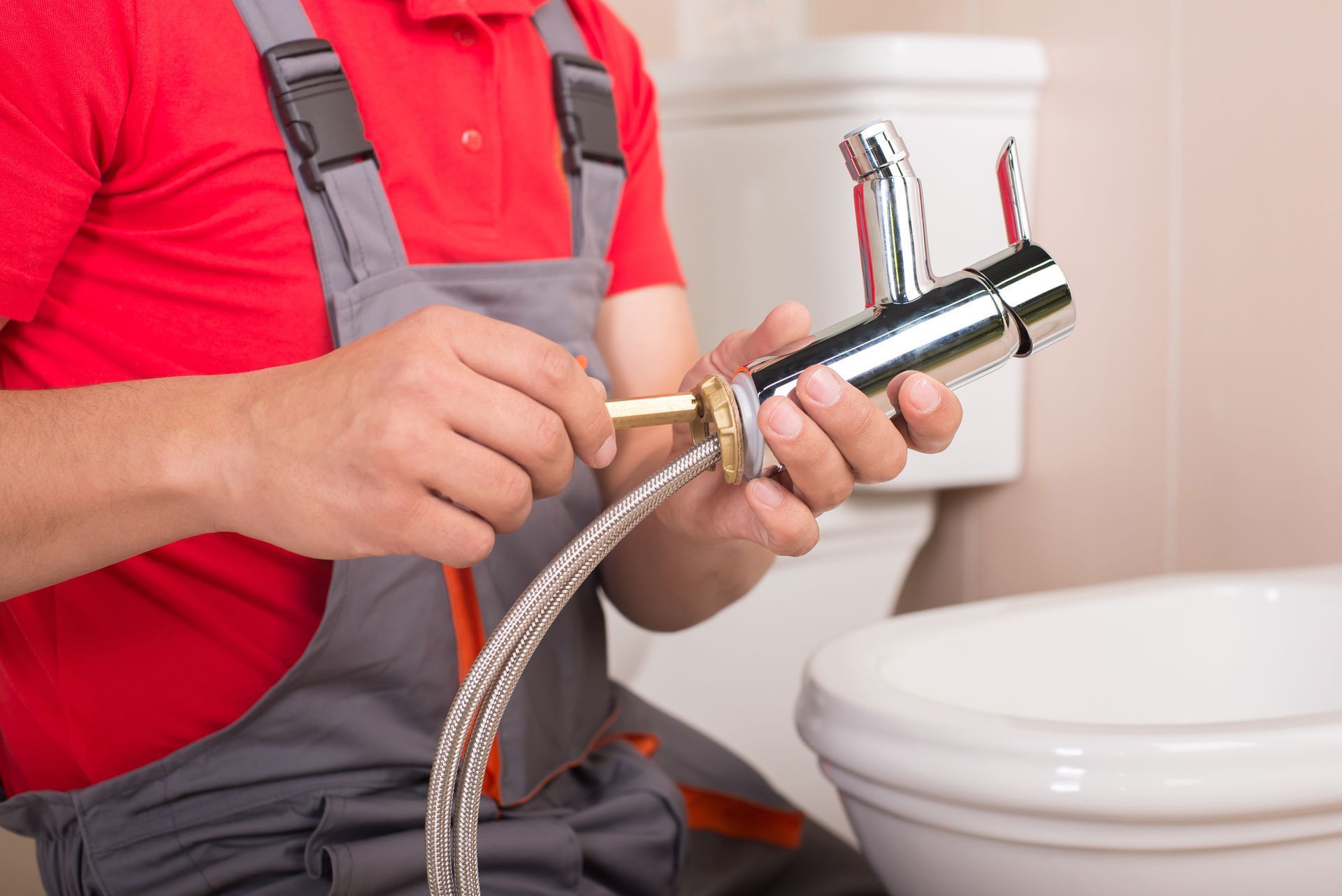 Plumber in red shirt and gray overalls installing a bathroom faucet near a toilet and sink.