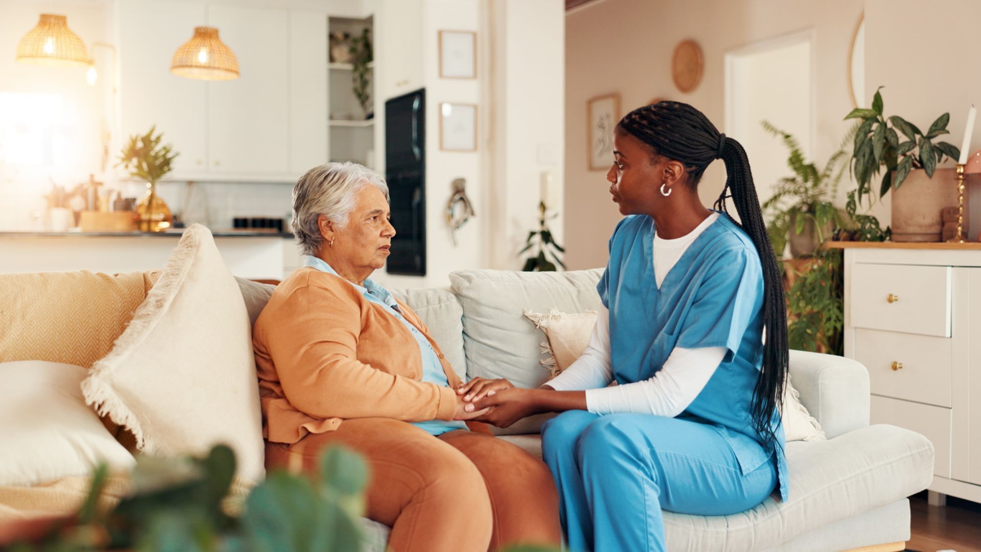 Caregiver in scrubs holds hands with a person on a sofa, inside a bright living room.