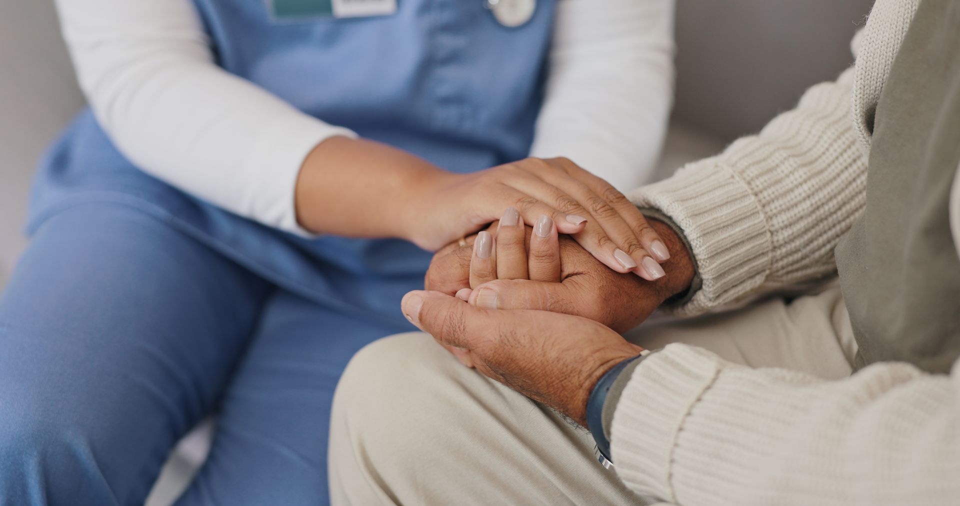 A healthcare worker in blue scrubs holds an elderly person's hands in comfort.