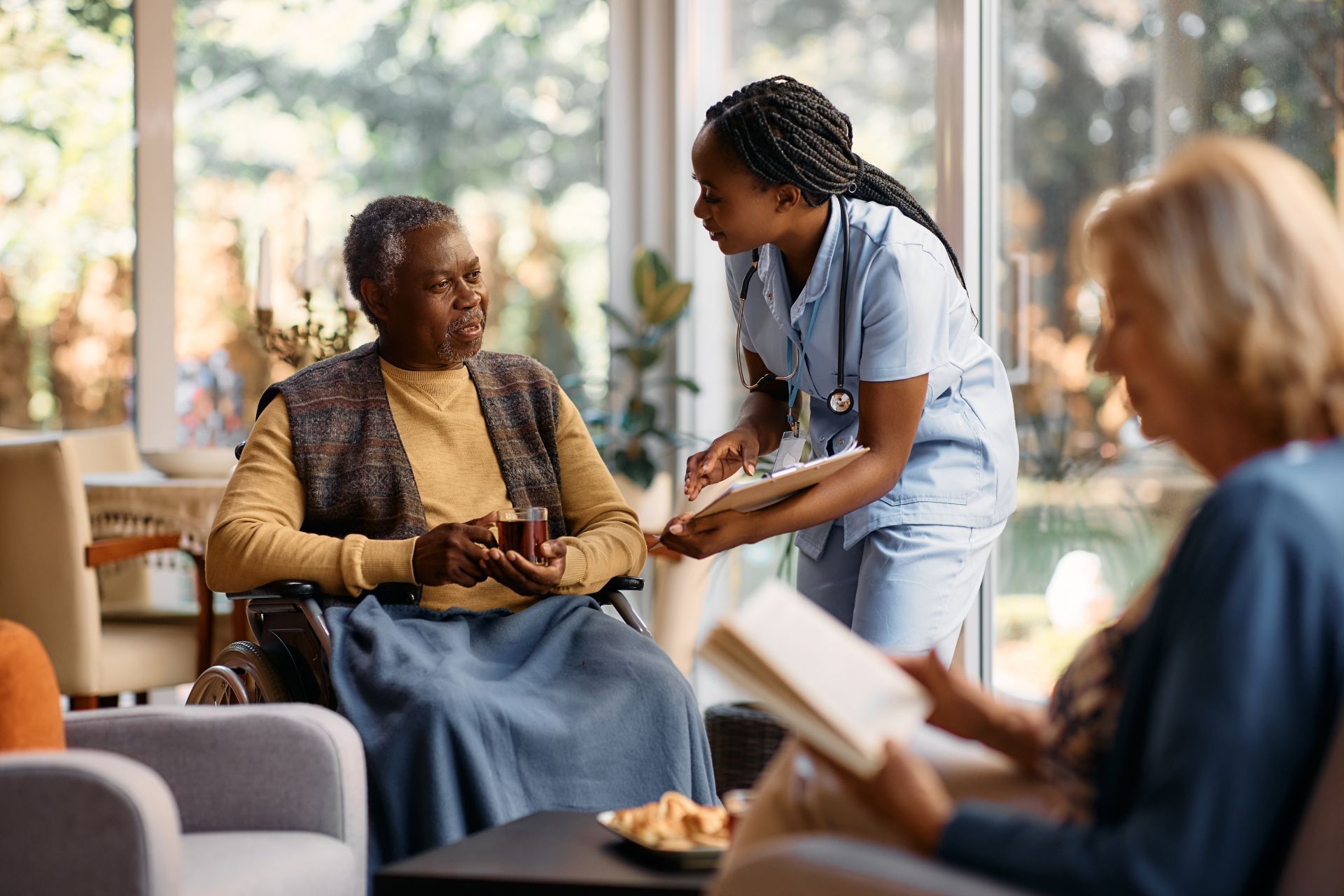 A caregiver in blue scrubs talks to a person in a wheelchair holding pills; another person reads nearby.