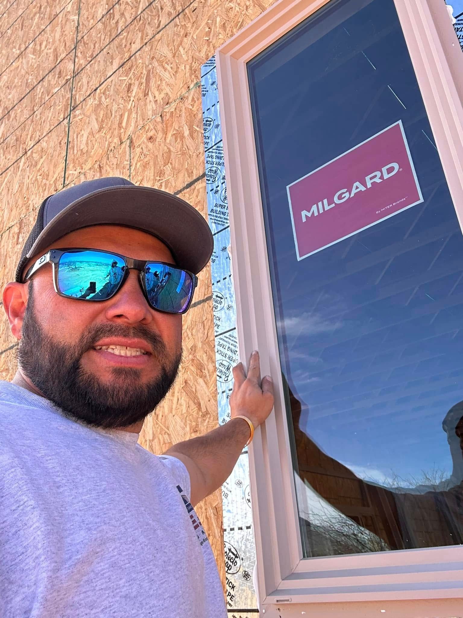 A man wearing sunglasses and a hat is standing next to a window.
