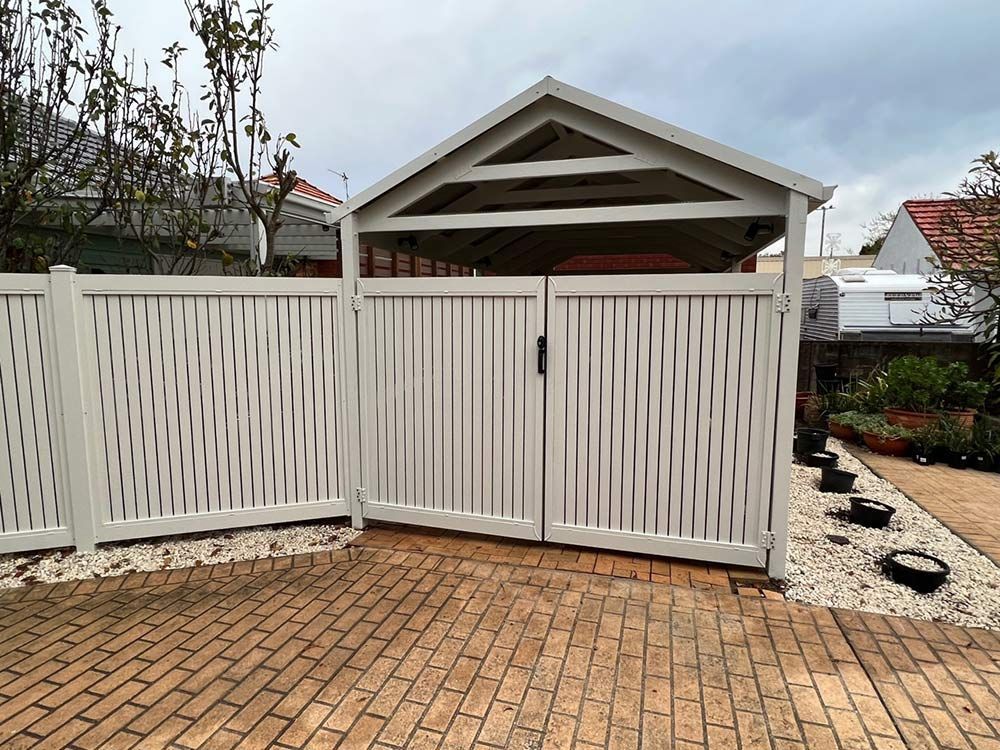 A White Fence Surrounds a Brick Driveway in Front of a House — Rearden Fencing Pty Ltd in Toronto, NSW