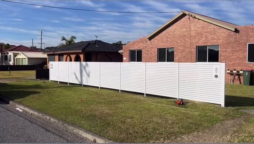 A White Fence is in Front of a Brick House — Rearden Fencing Pty Ltd in Toronto, NSW
