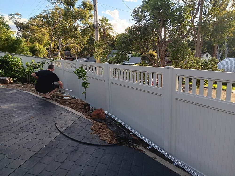 A Man is Squatting Down Next to a White Fence — Rearden Fencing Pty Ltd in Toronto, NSW