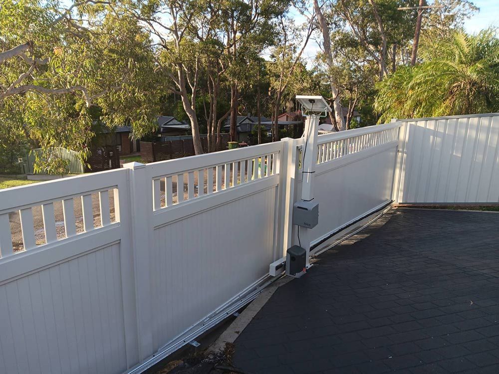 A White Fence is Surrounding a Driveway With Trees in the Background — Rearden Fencing Pty Ltd in Toronto, NSW