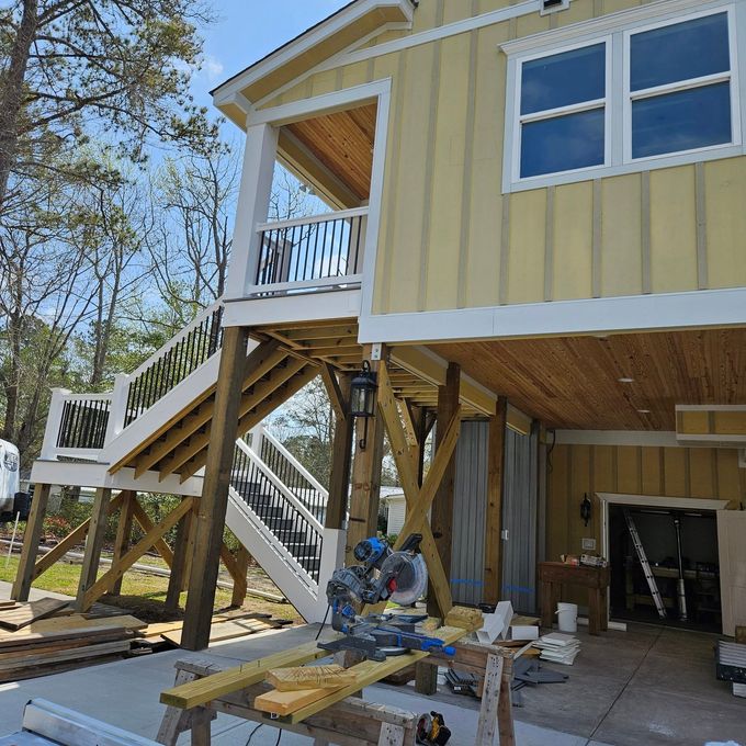 A craftsman cuts wood at a construction site with a yellow-sided raised house and stairs.