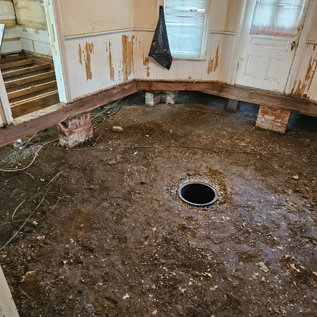 Room under construction; dirt floor, exposed beams, brick supports, dark drain, peeling paint, stairs.