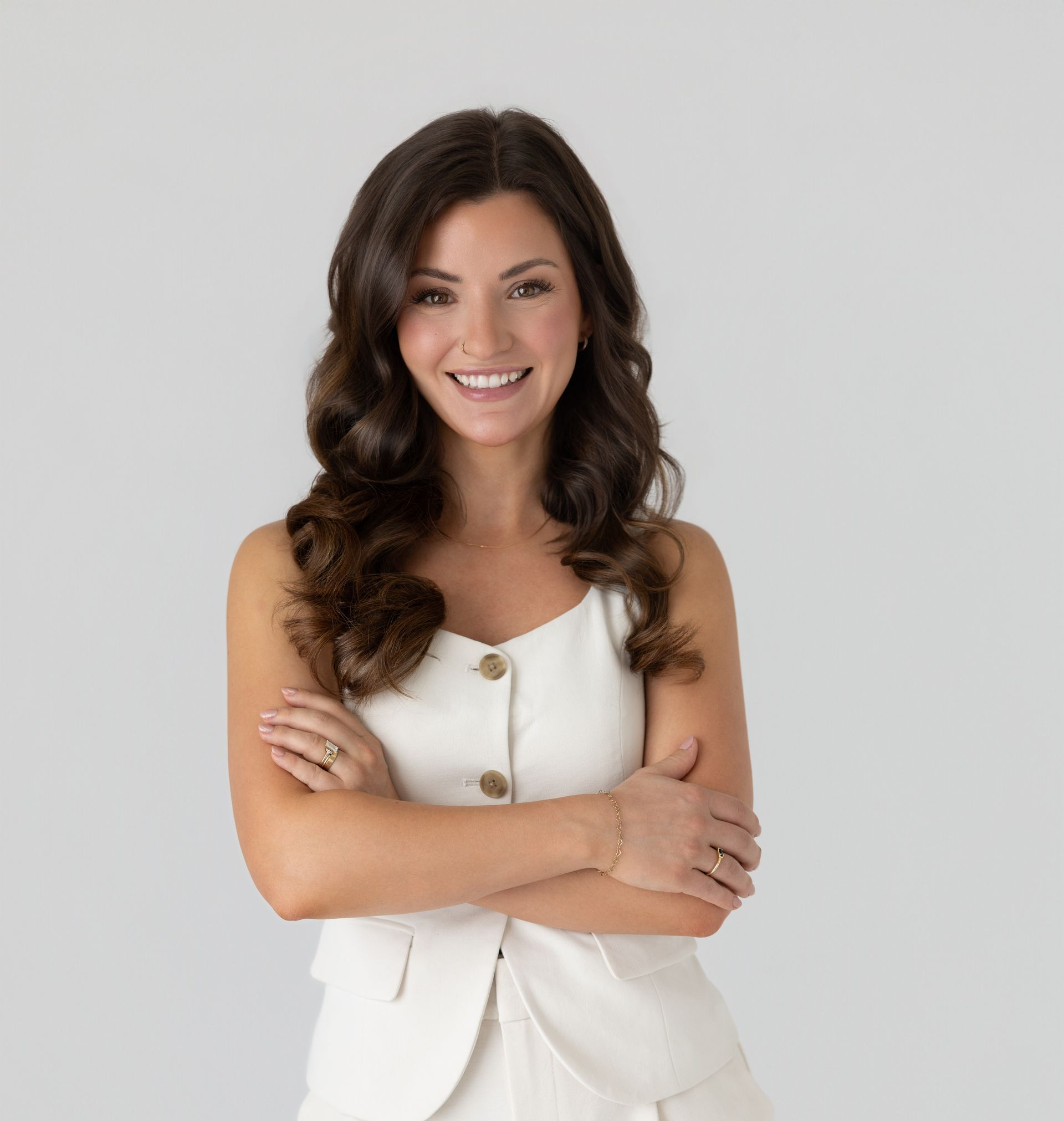Smiling woman with long dark hair, arms crossed, wearing a white sleeveless outfit against a light gray background