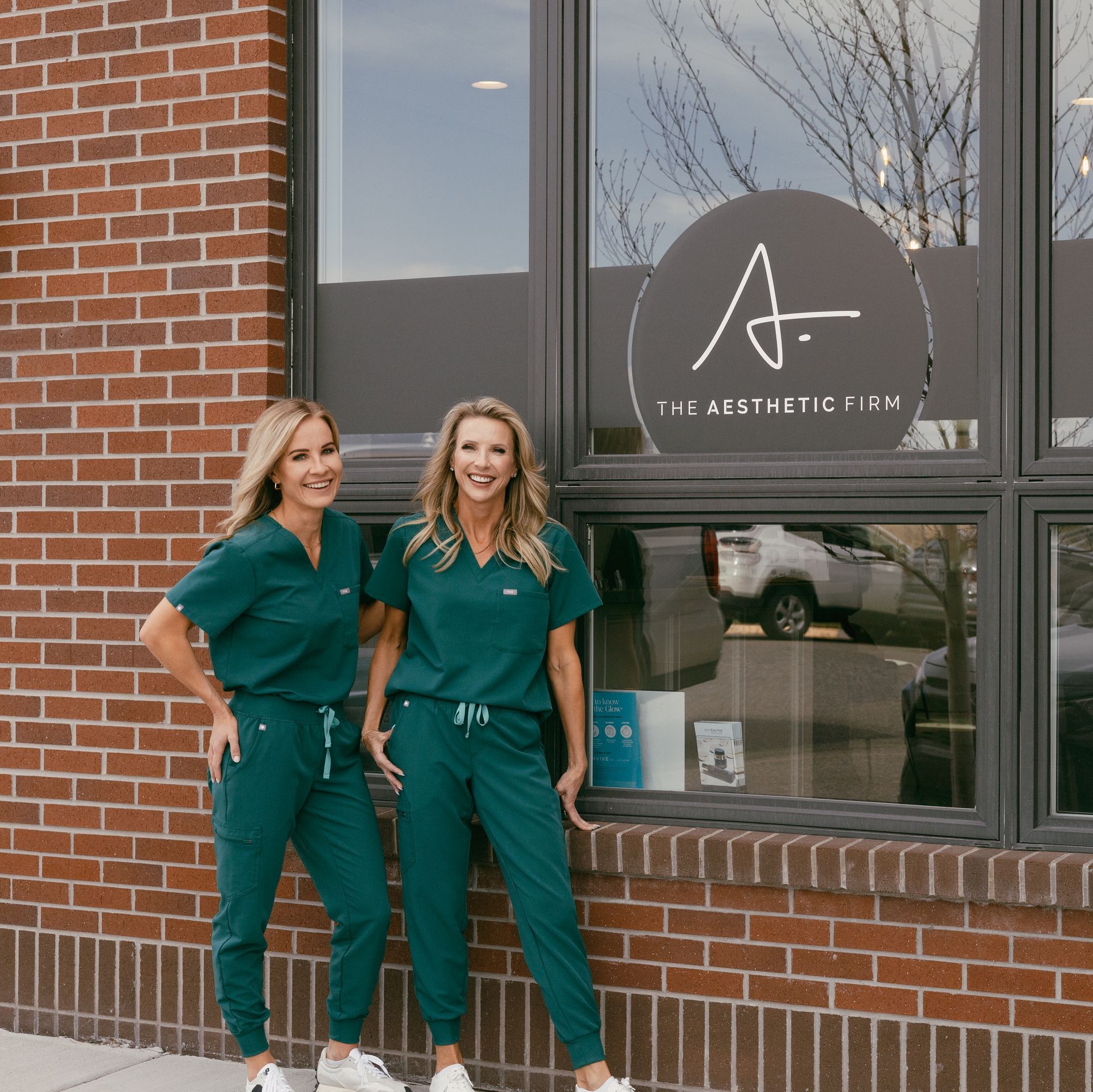 Two women in green scrubs are standing in front of a brick building.