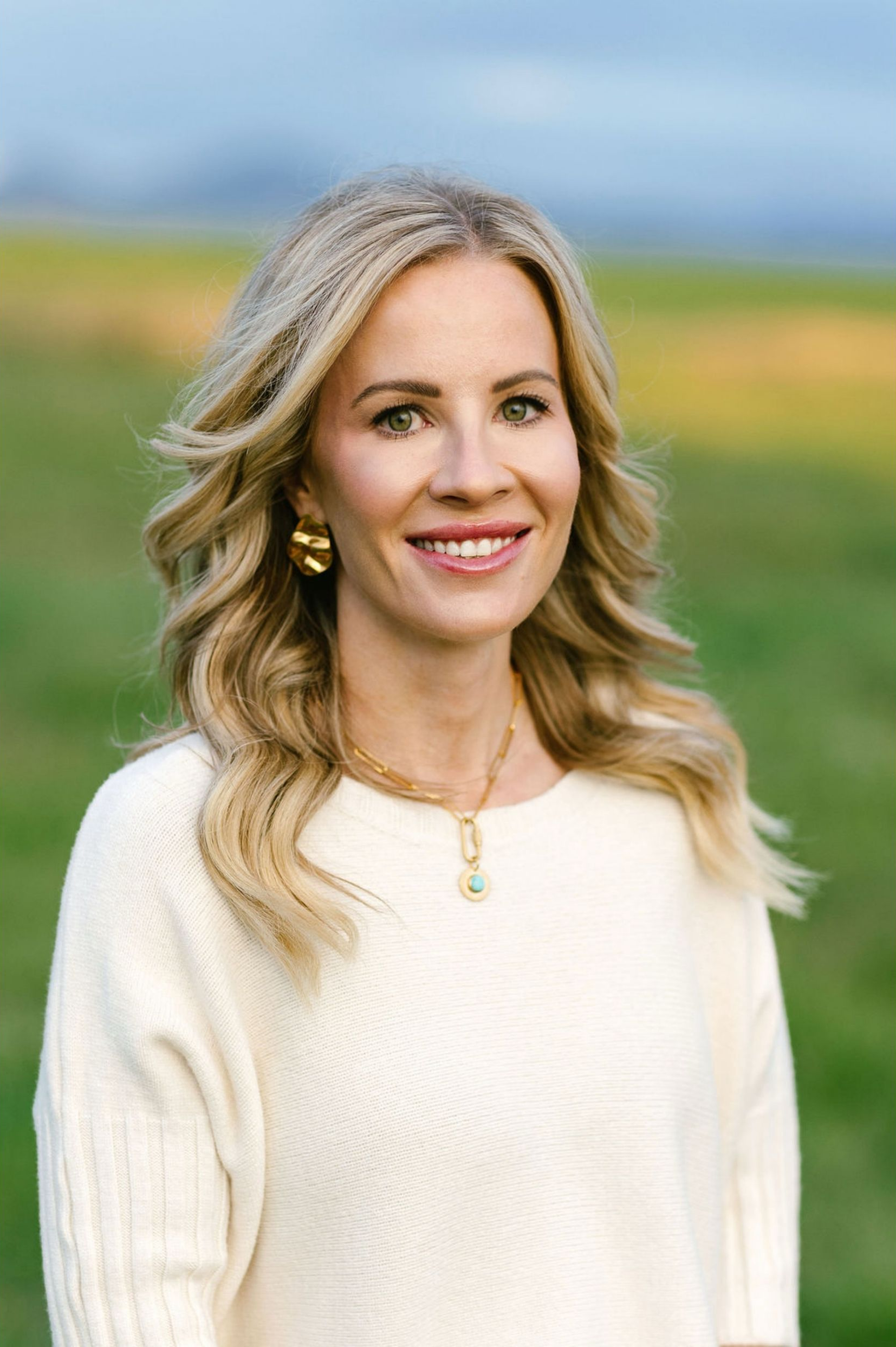 Smiling woman in beige top and white pants seated on a cushioned bench in a bright room