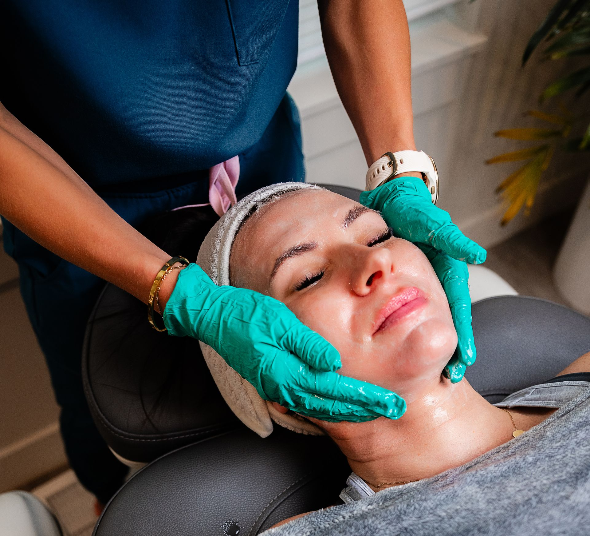Esthetician giving a facial massage to a reclining client in green gloves and a headband.