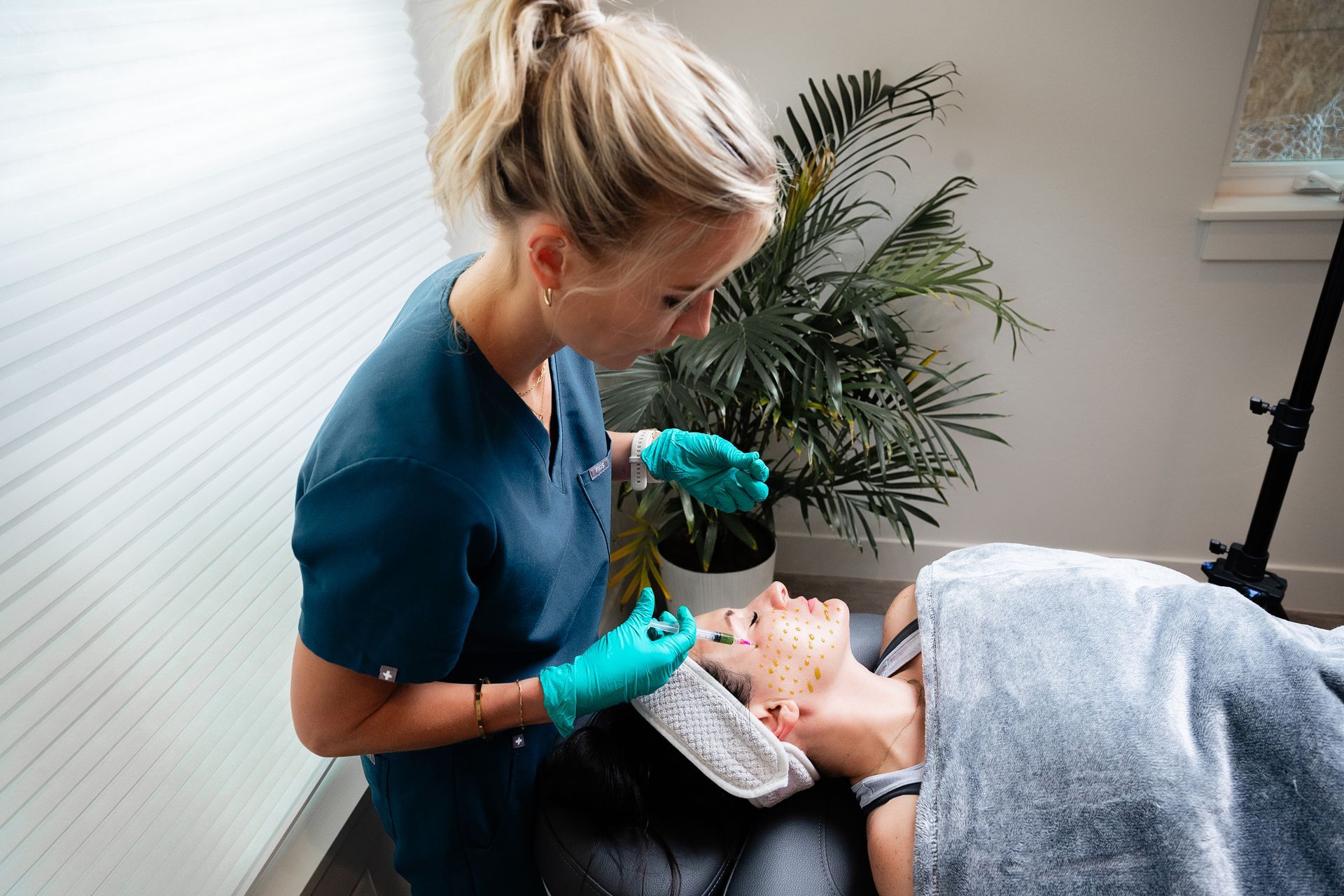 Esthetician wearing teal gloves applies a facial treatment to a client lying under a gray towel.